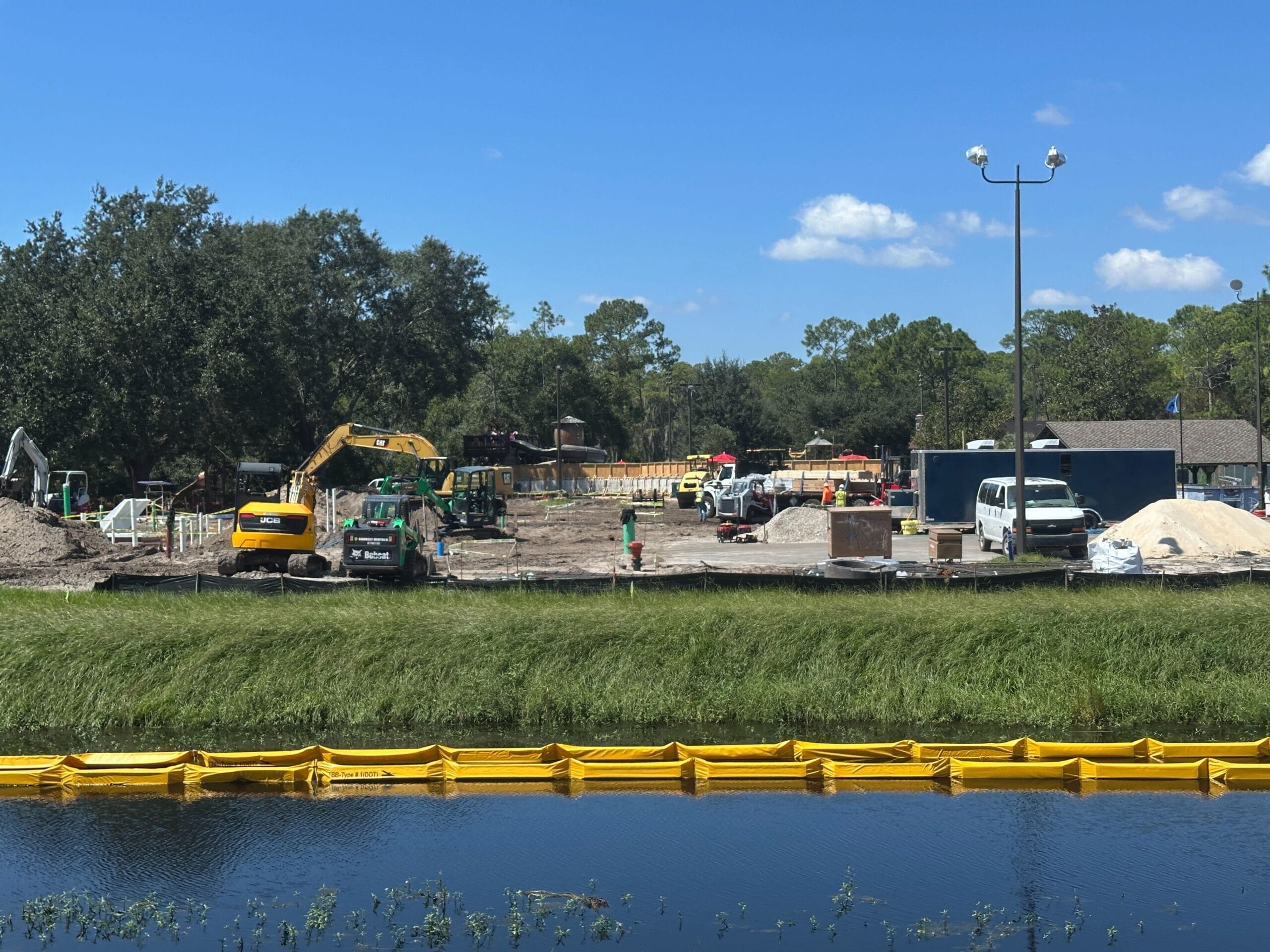 Construction site with machinery, workers, and vehicles near a pond with a yellow barrier and trees in the background.