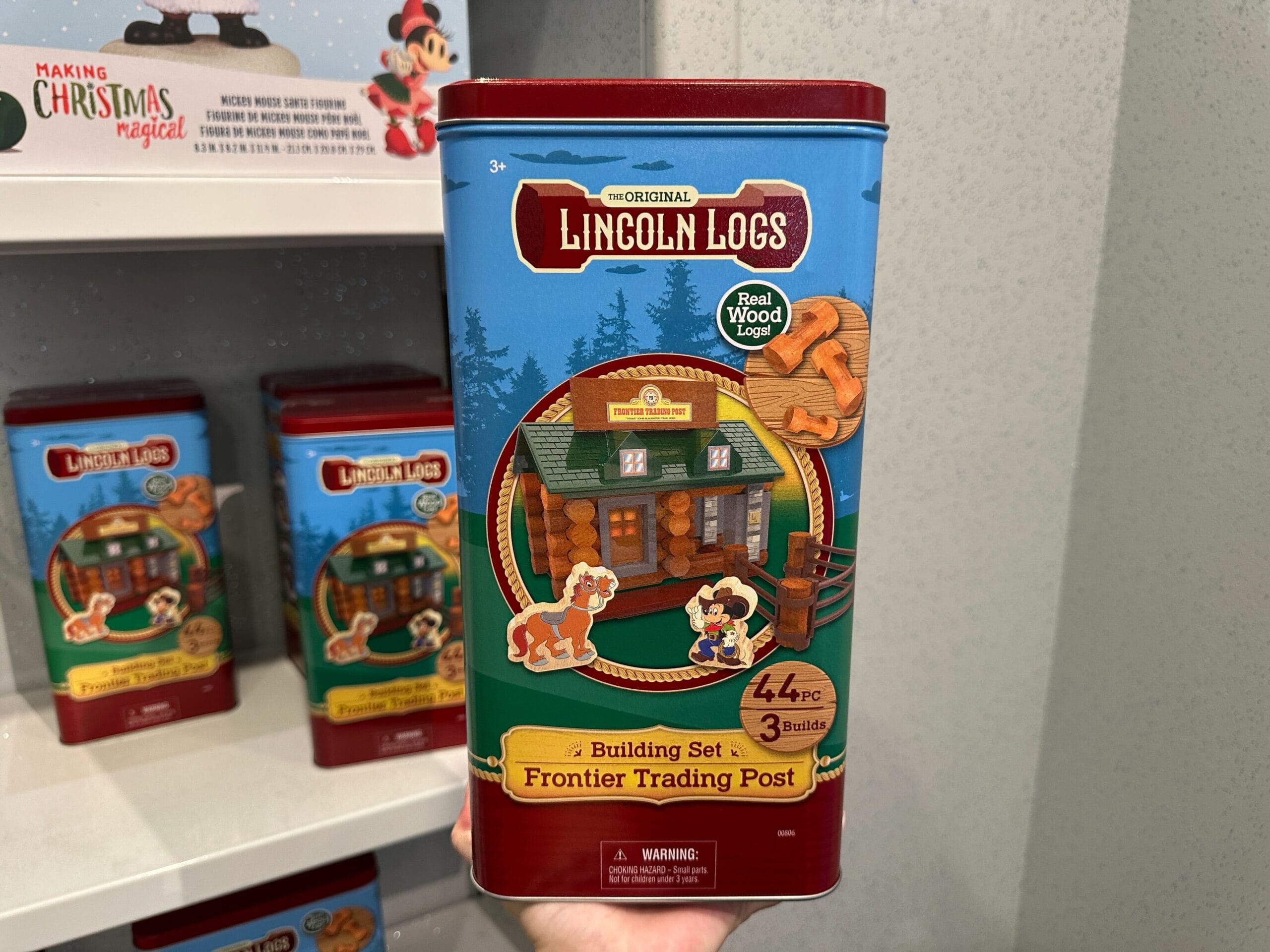 A hand holds a Lincoln Logs Frontier Trading Post building set tin with colorful illustrations on the front.