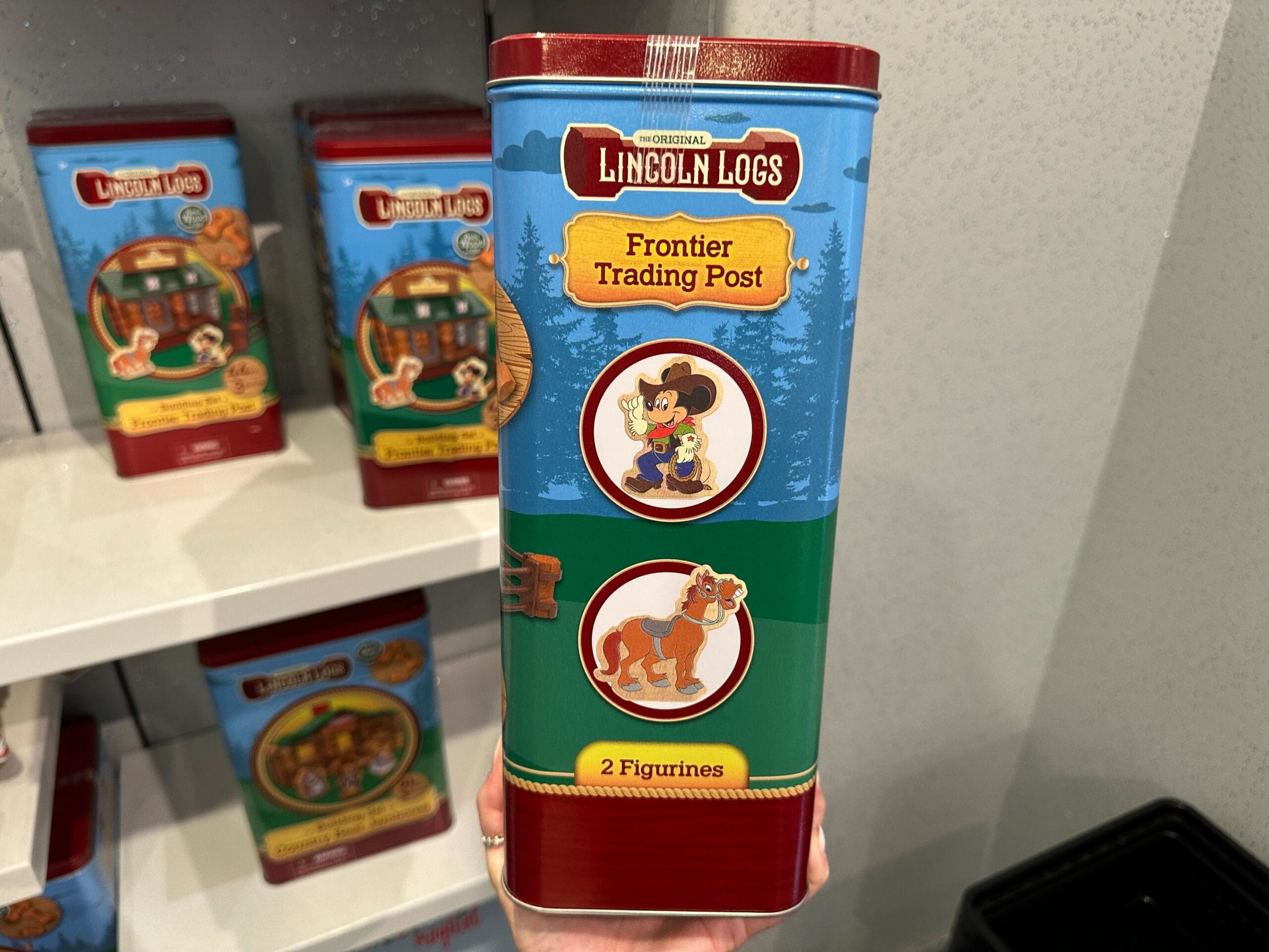 A hand holds a Lincoln Logs Frontier Trading Post tin with cartoon cowboy and horse images and "2 Figurines" text.