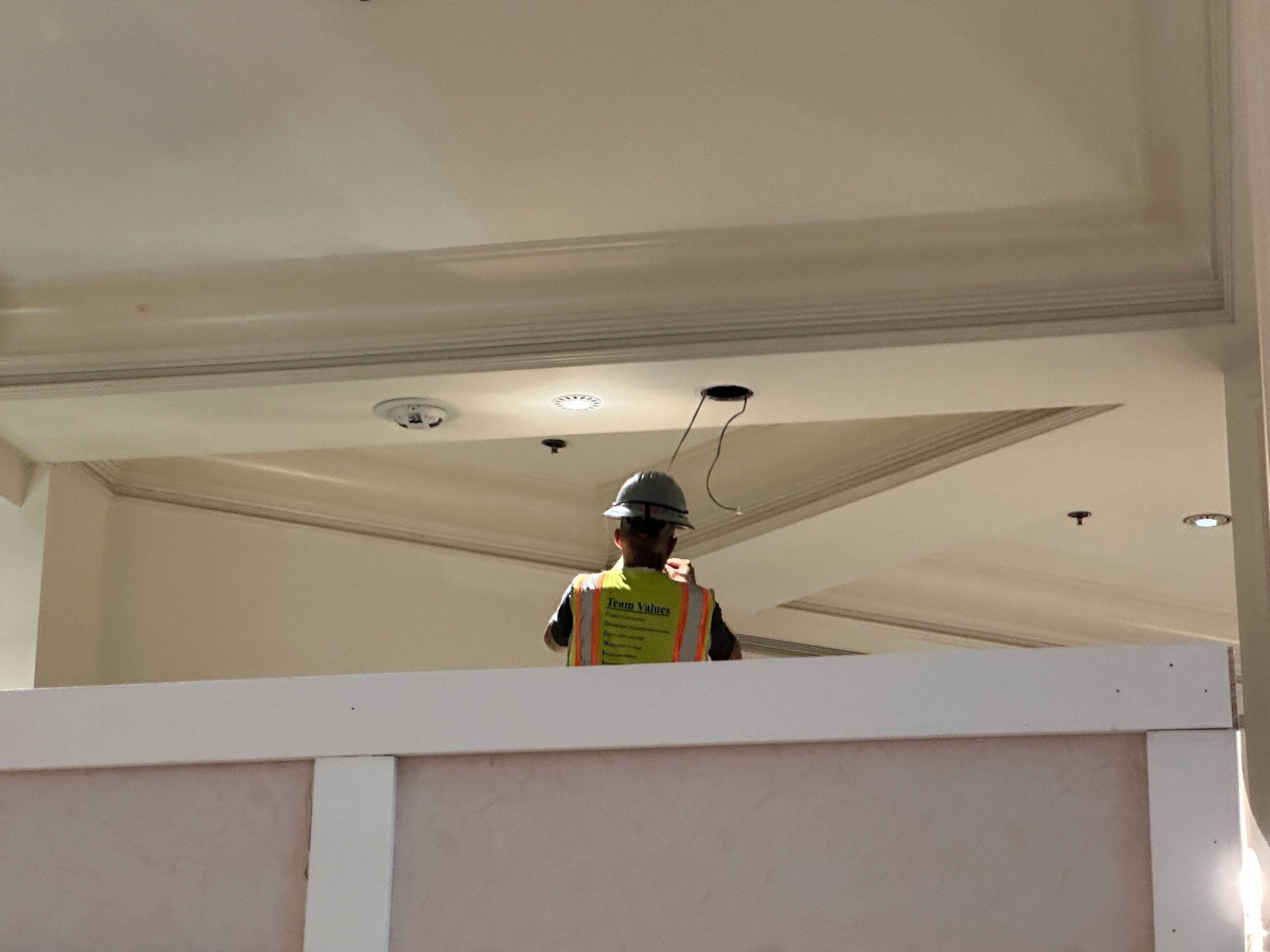 Construction worker in a hard hat and vest stands by a ceiling with exposed wiring, partially obscured by a partition.