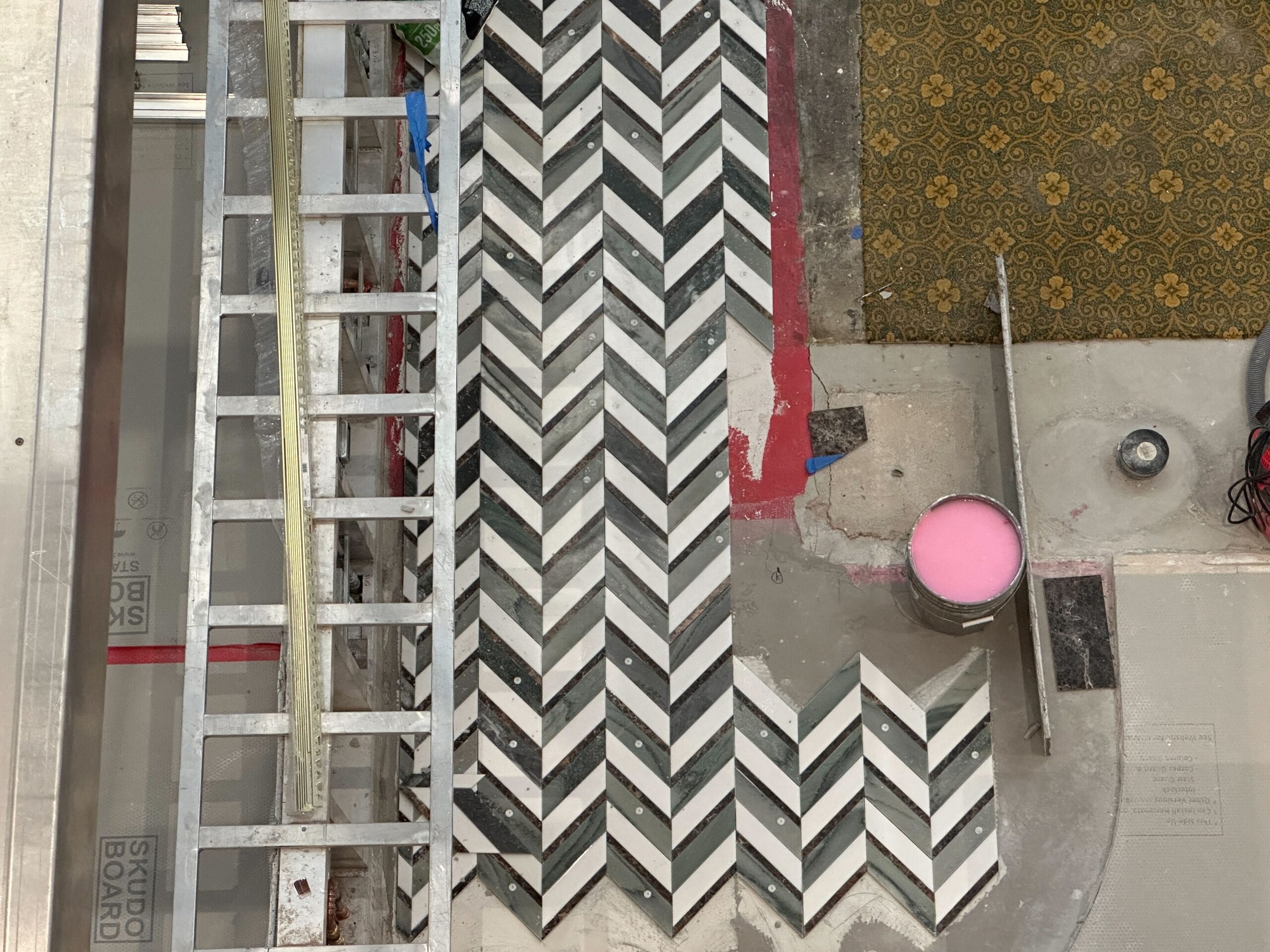 Overhead view of patterned tiles being installed, with a ladder, pink bucket, tools, and carpet nearby.