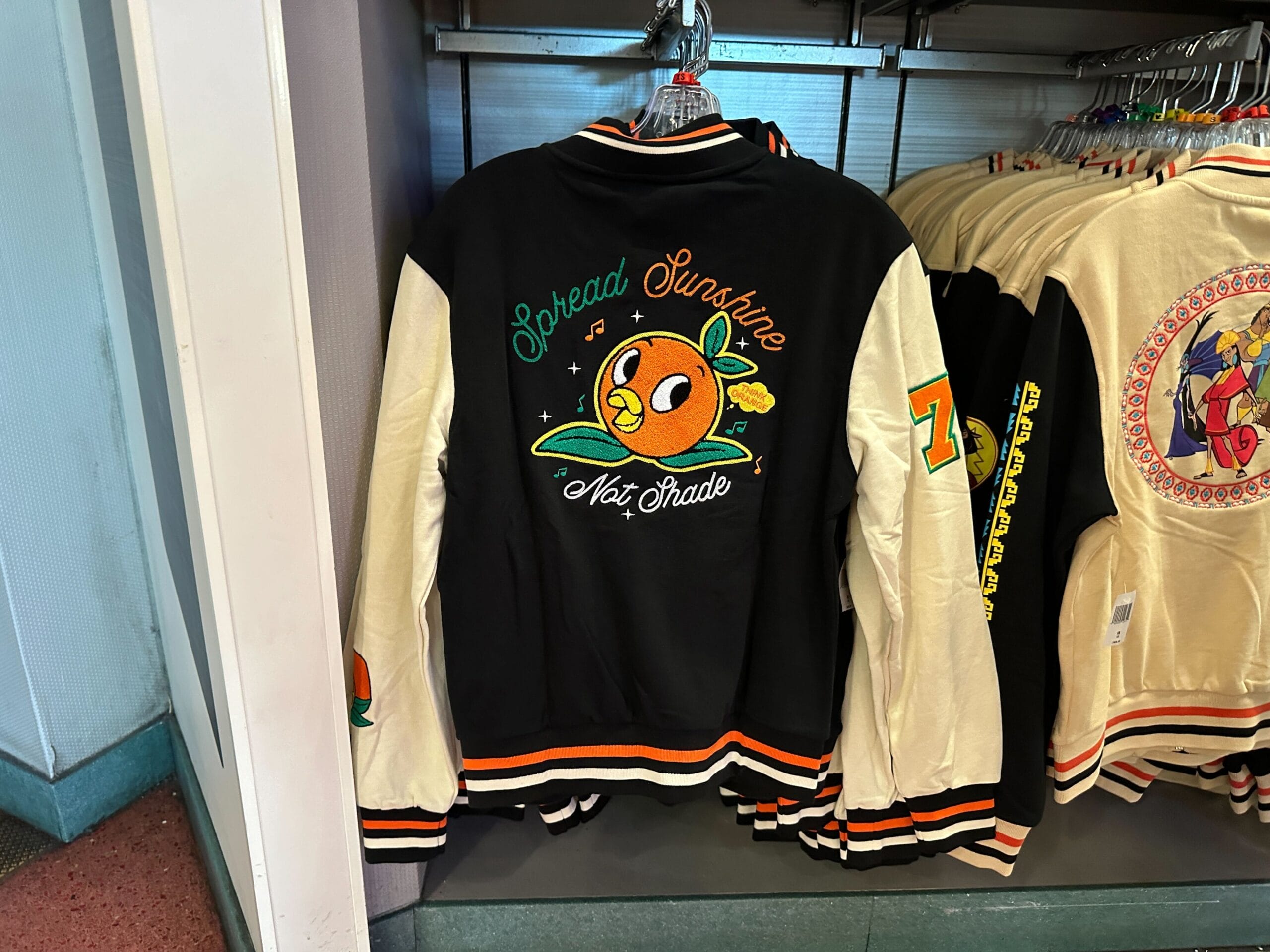 An Orange Bird varsity jacket with "Spread Sunshine Not Shade" hangs on a rack at Walt Disney World.