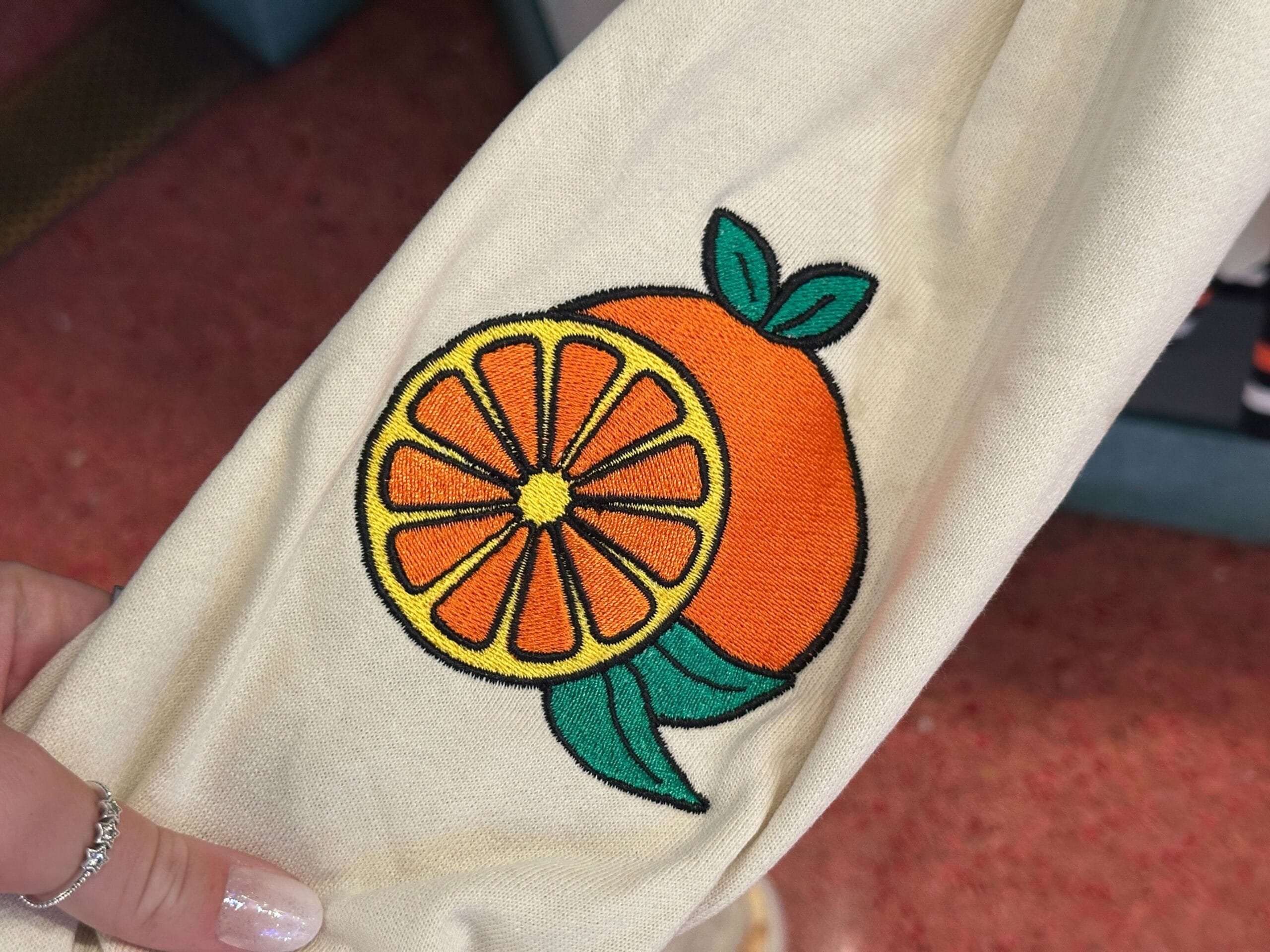 A hand holds fabric with an embroidered Orange Bird fruit and segment design, accented with green leaves.
