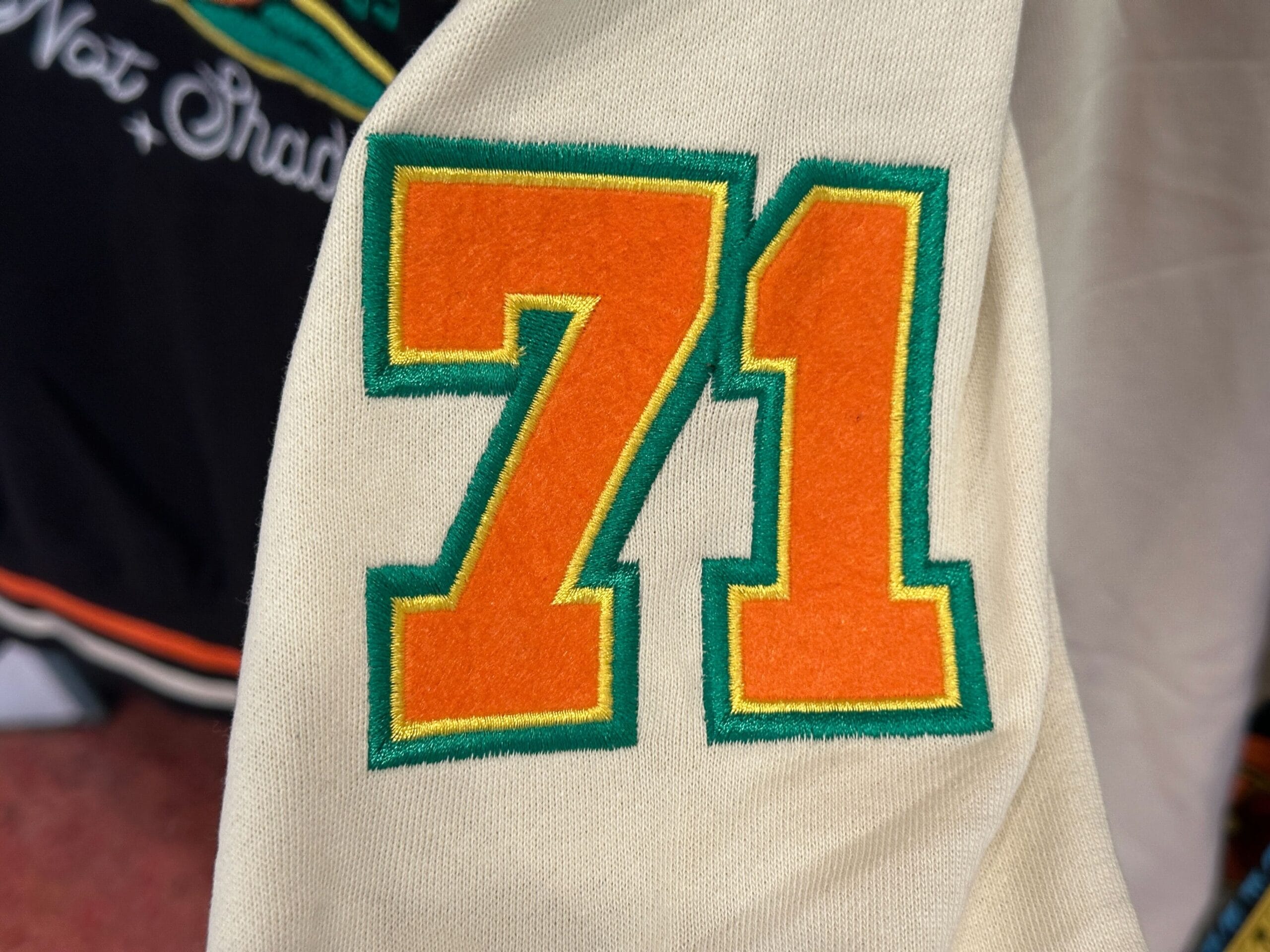 Orange and yellow number 71 with a green outline, inspired by Disney Letterman Jackets, embroidered on cream fabric.