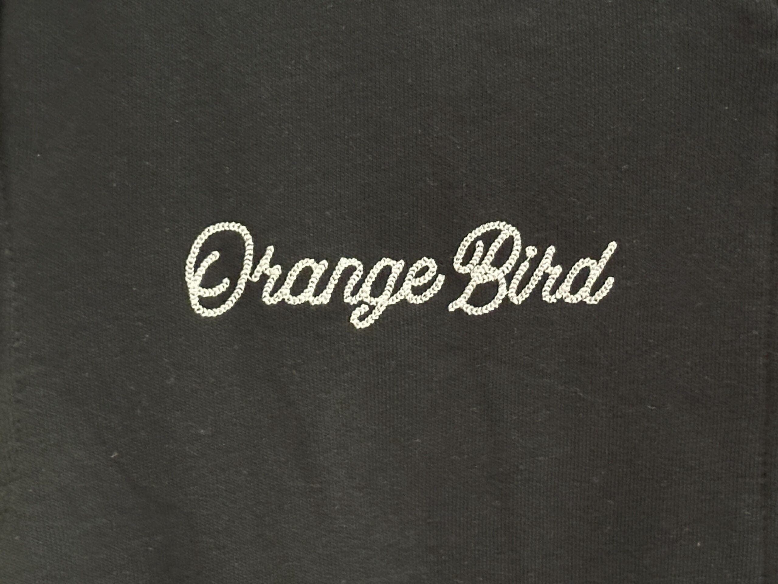 Black fabric with "Orange Bird" in a white, dotted font—perfect for Disney Letterman Jackets fans.