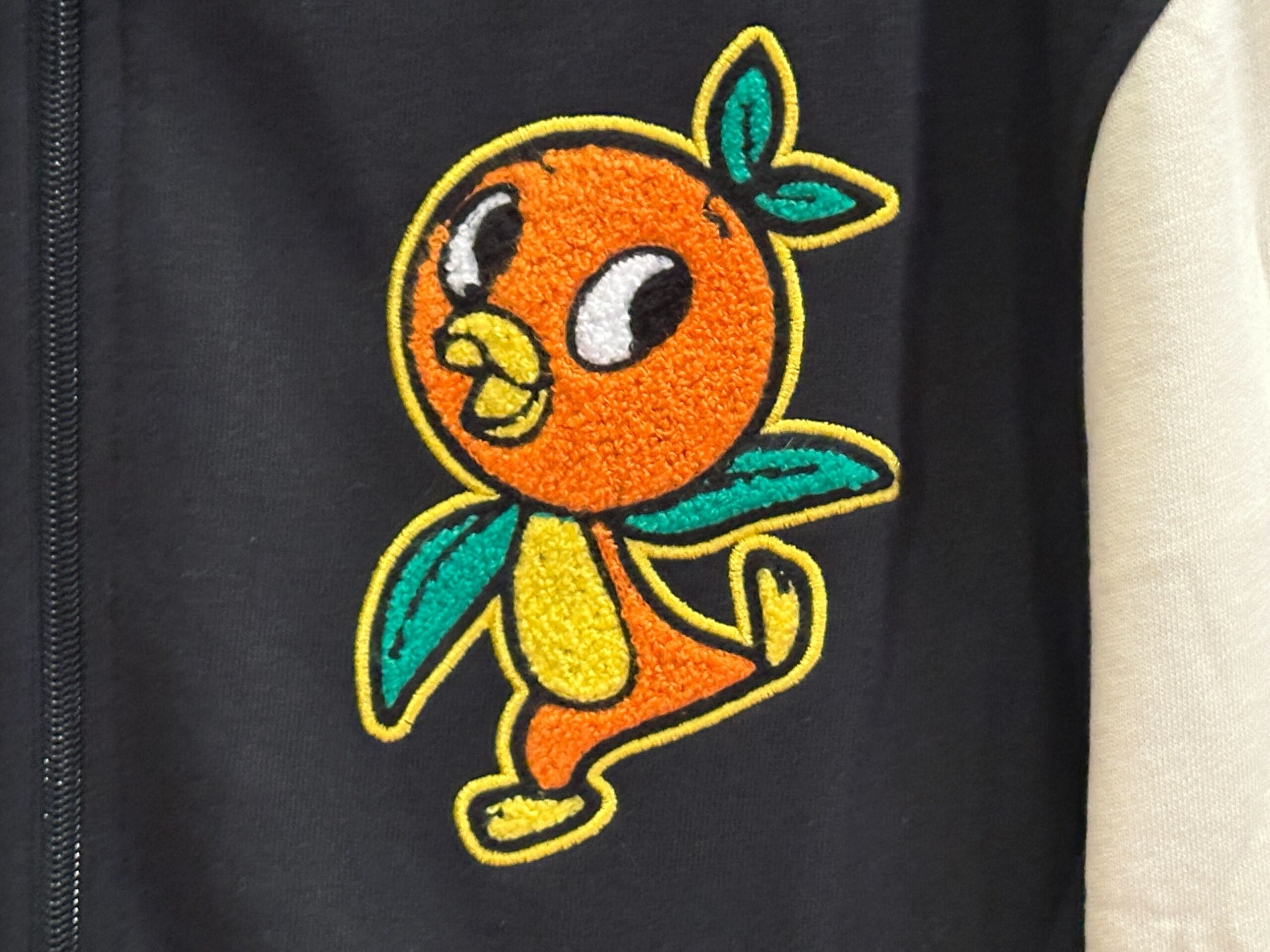 Colorful cartoon Orange Bird patch with green leaves on black fabric, next to a light-colored Disney Letterman Jacket sleeve.