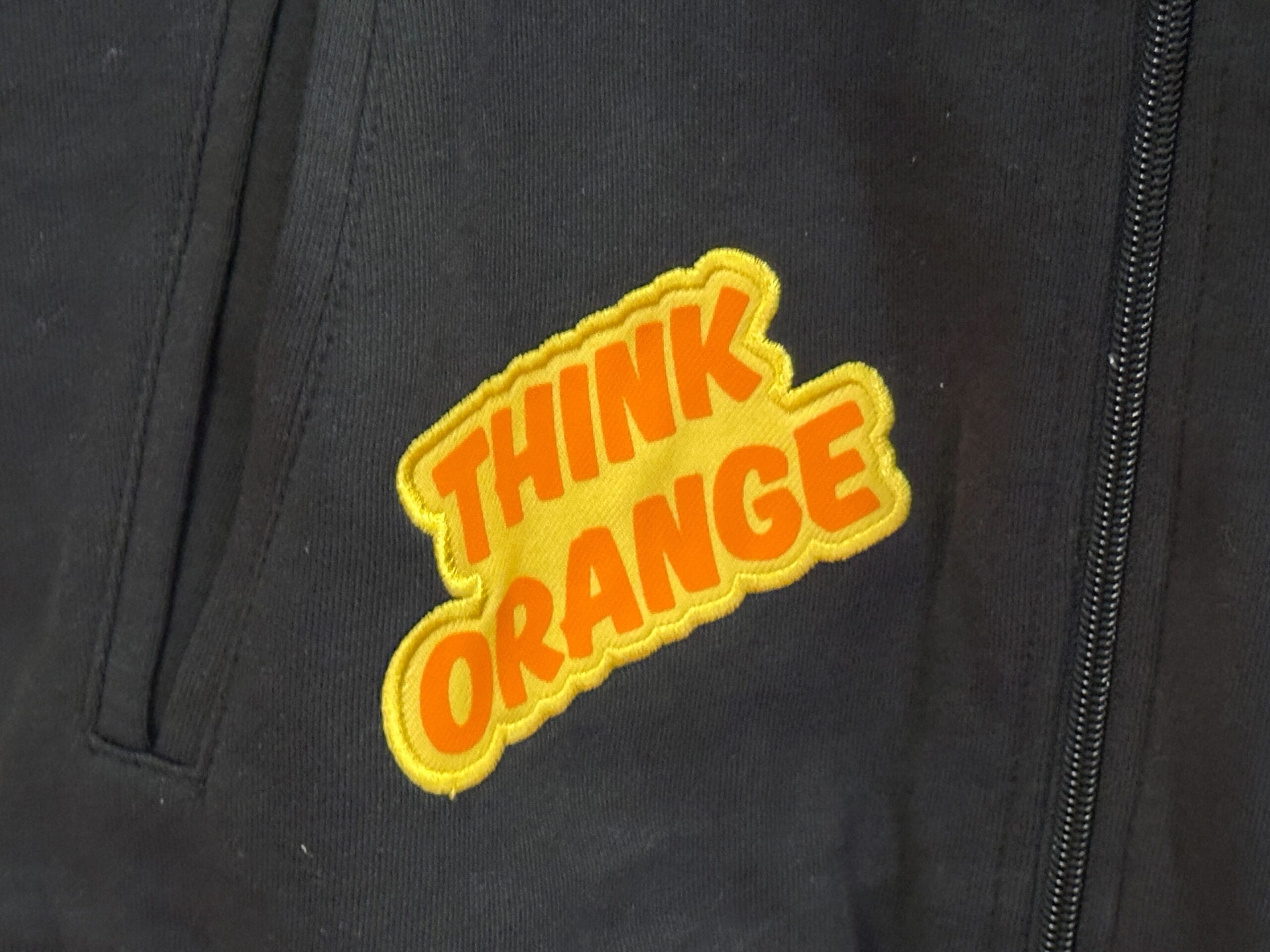 A black garment with a "THINK ORANGE" patch near the zipper, inspired by Orange Bird from Walt Disney World.