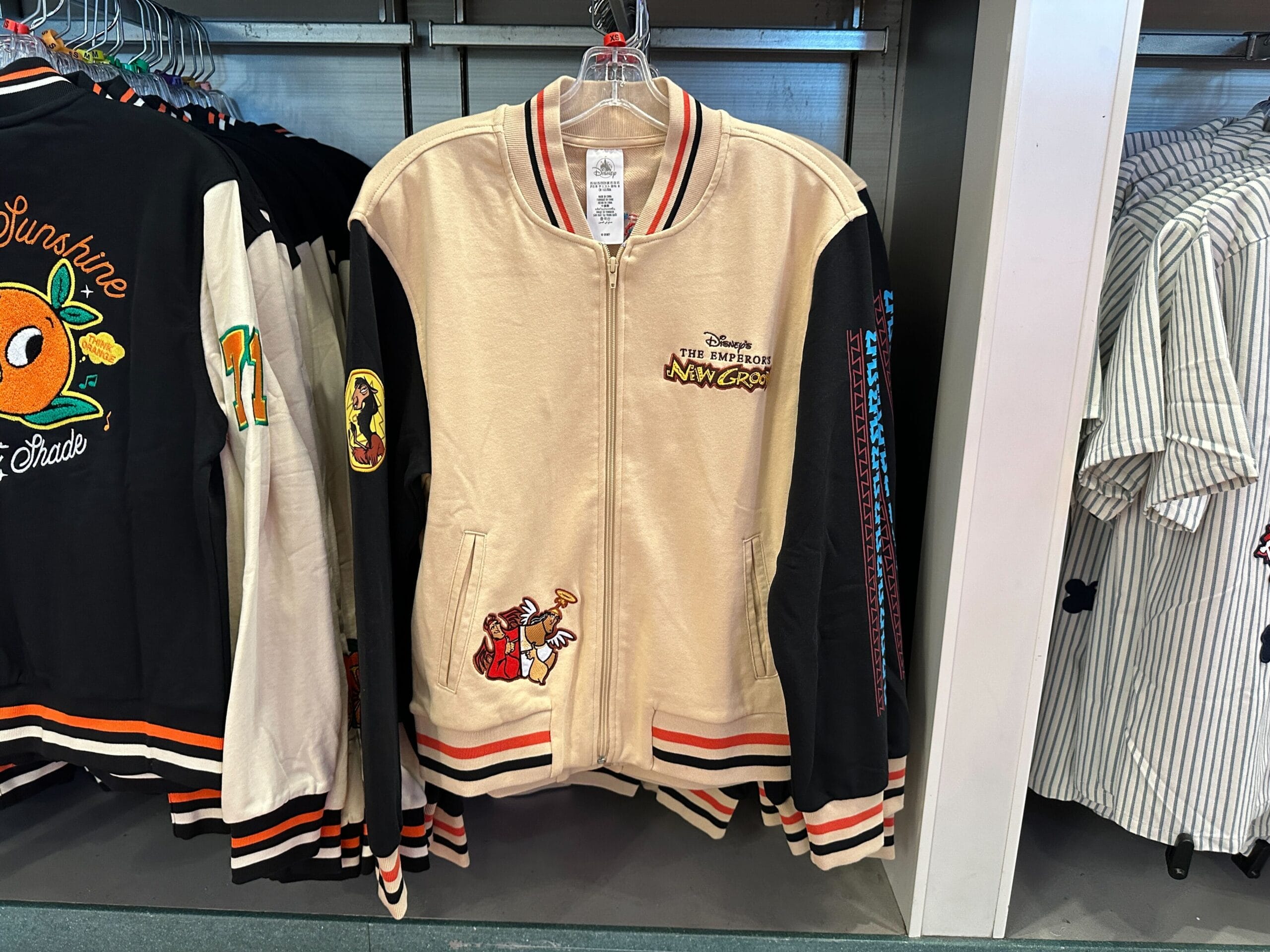 Beige and black Disney Letterman Jacket featuring "The Emperor's New Groove" logo and cartoon designs on front and sleeve.