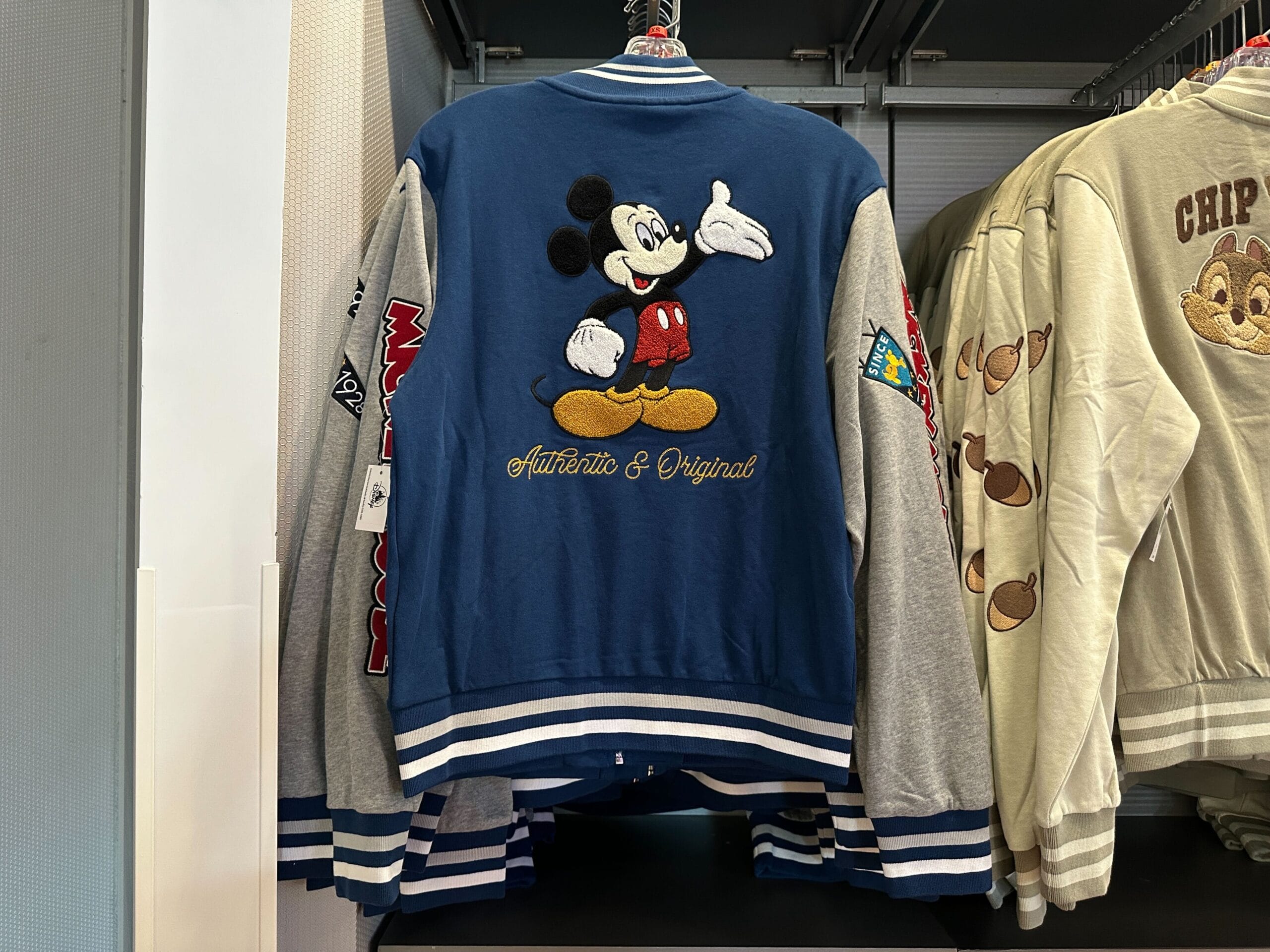 Blue and gray Disney Letterman jacket featuring Mickey Mouse and “Authentic & Original” text hanging on a store rack.