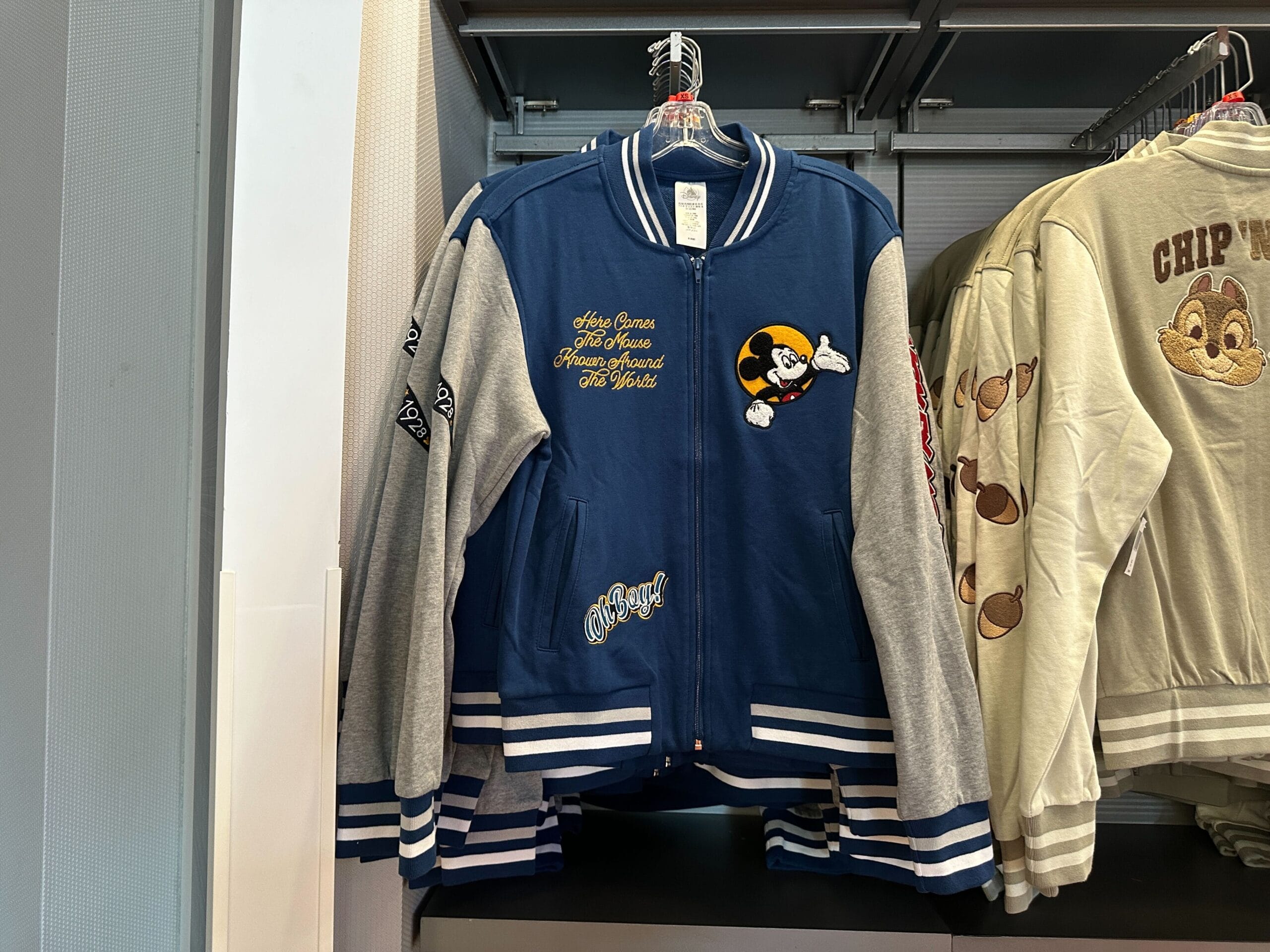 Blue and gray Disney Letterman Jacket featuring a Mickey Mouse patch, hanging on a clothing rack in Walt Disney World.