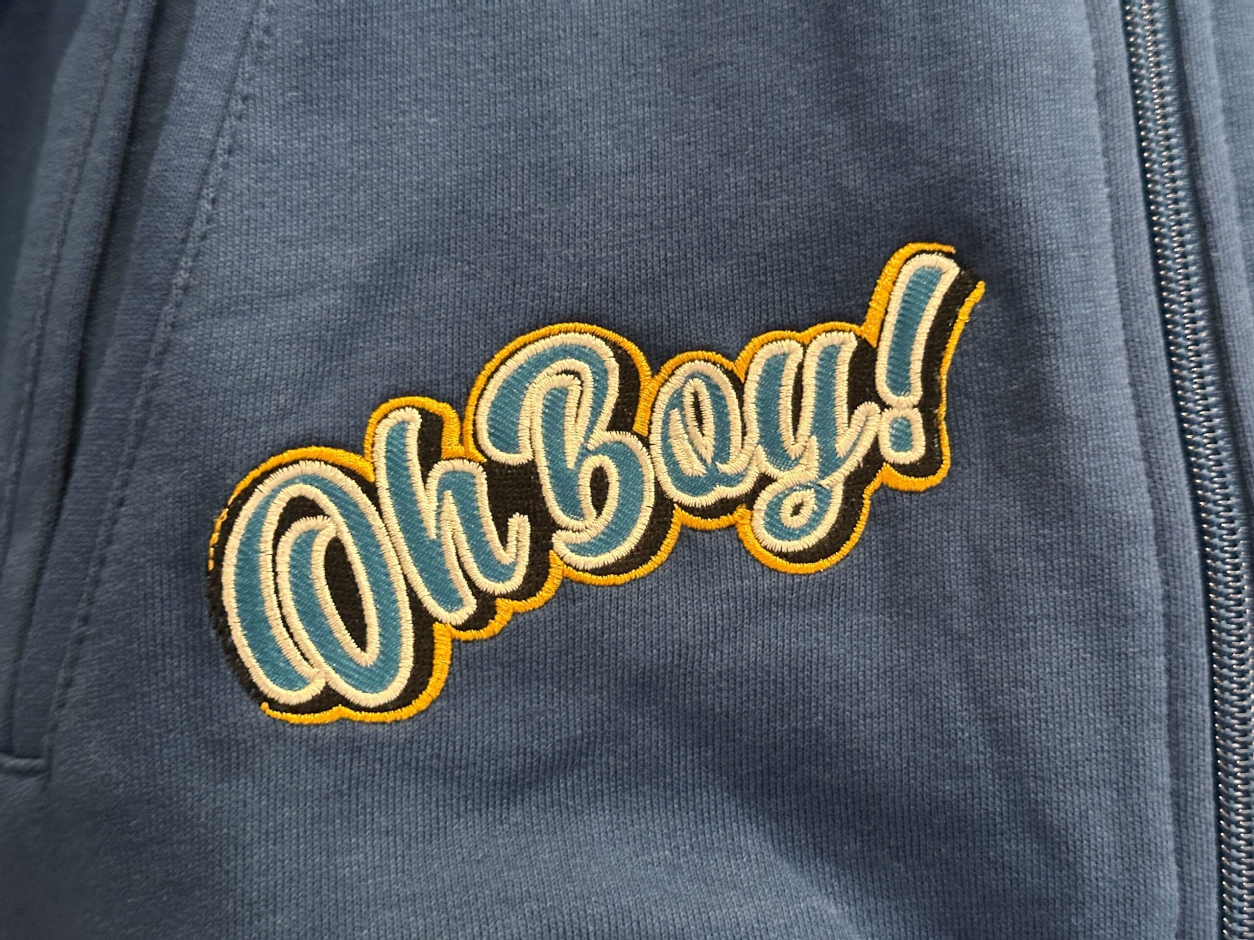 Blue fabric with embroidered "Oh Boy!" text, reminiscent of Disney Letterman Jackets, next to a zipper.