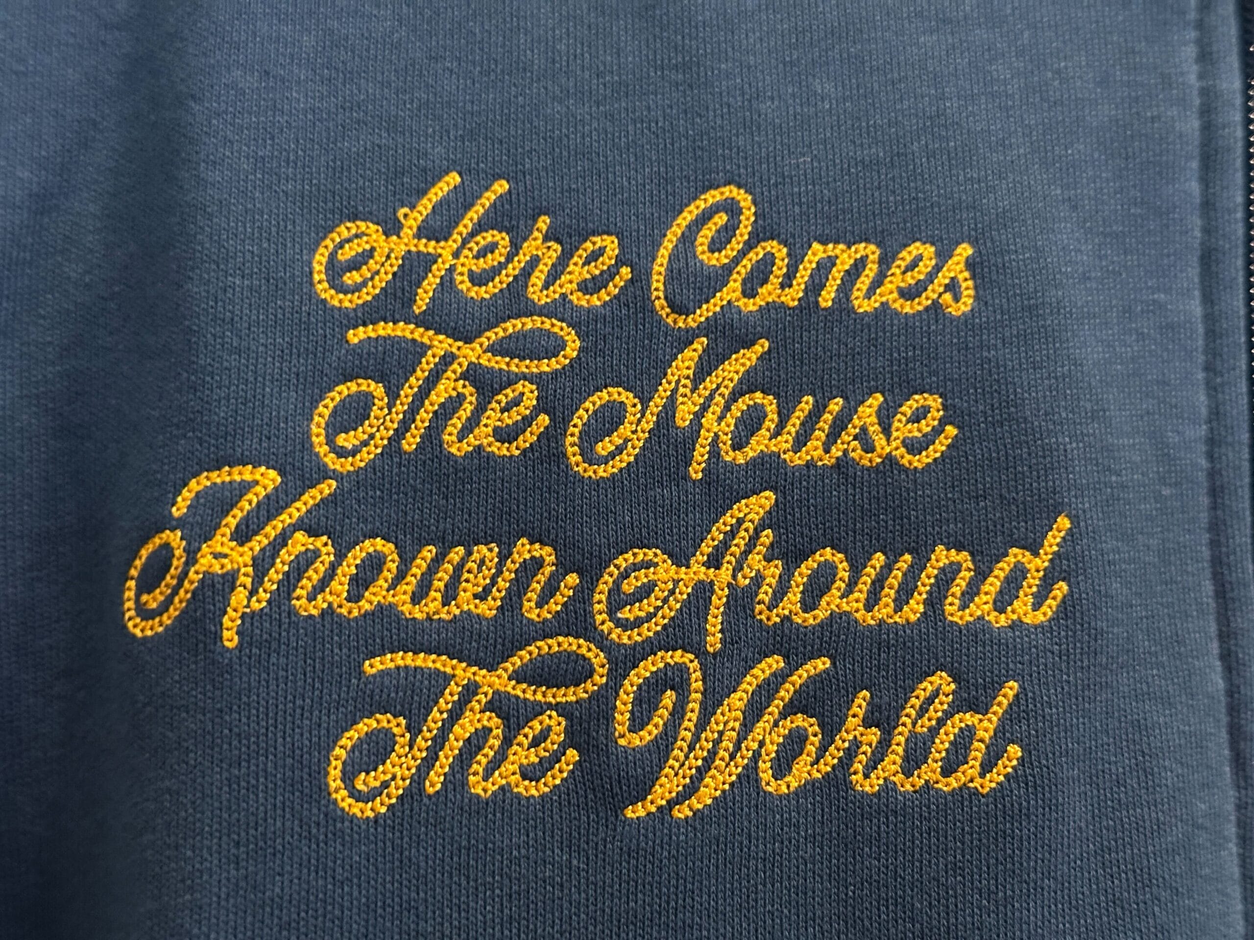 Yellow script text on dark fabric reads, "Here Comes The Mouse Known Around The World" at Walt Disney World.
