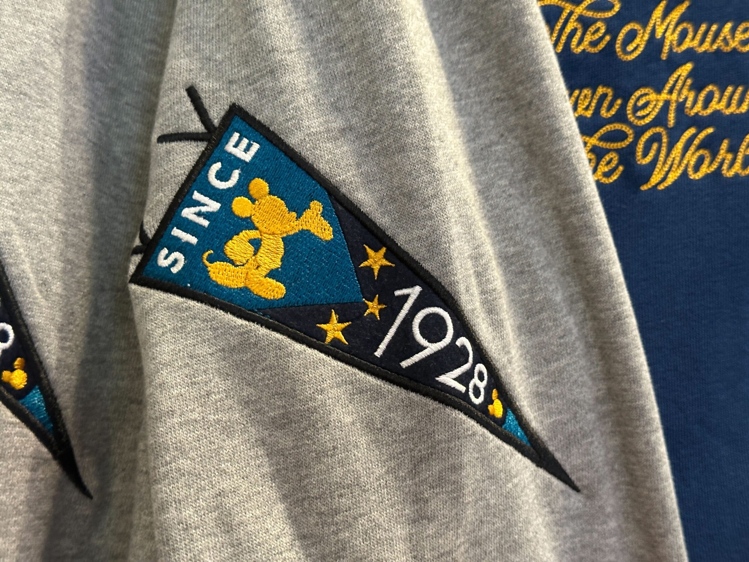 Gray shirt with a pennant patch showing Mickey Mouse, stars, and "Since 1928"—Disney Letterman Jackets style.