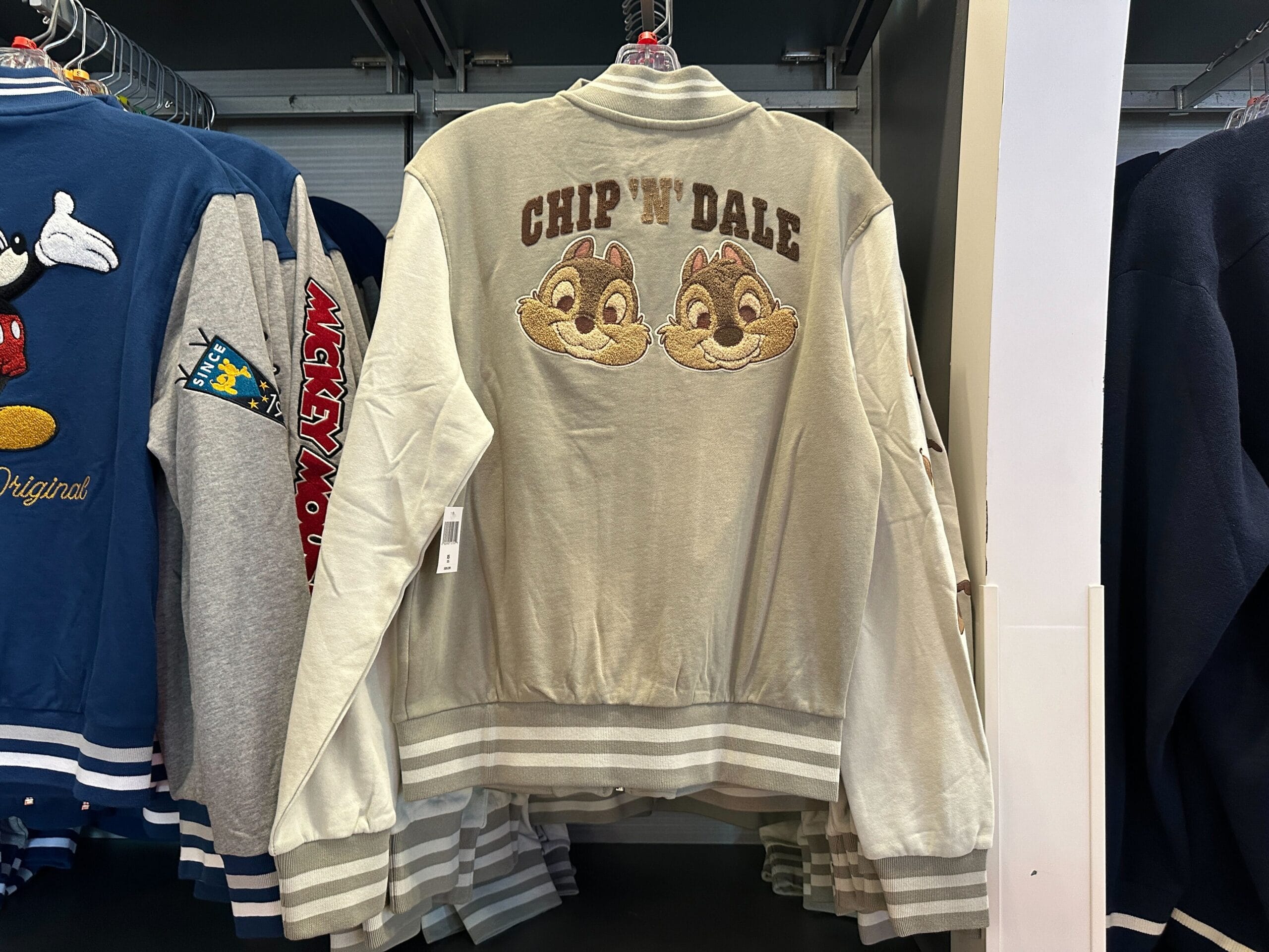 A light gray Disney Letterman Jacket with embroidered Chip 'n' Dale faces and text, hanging on a rack.