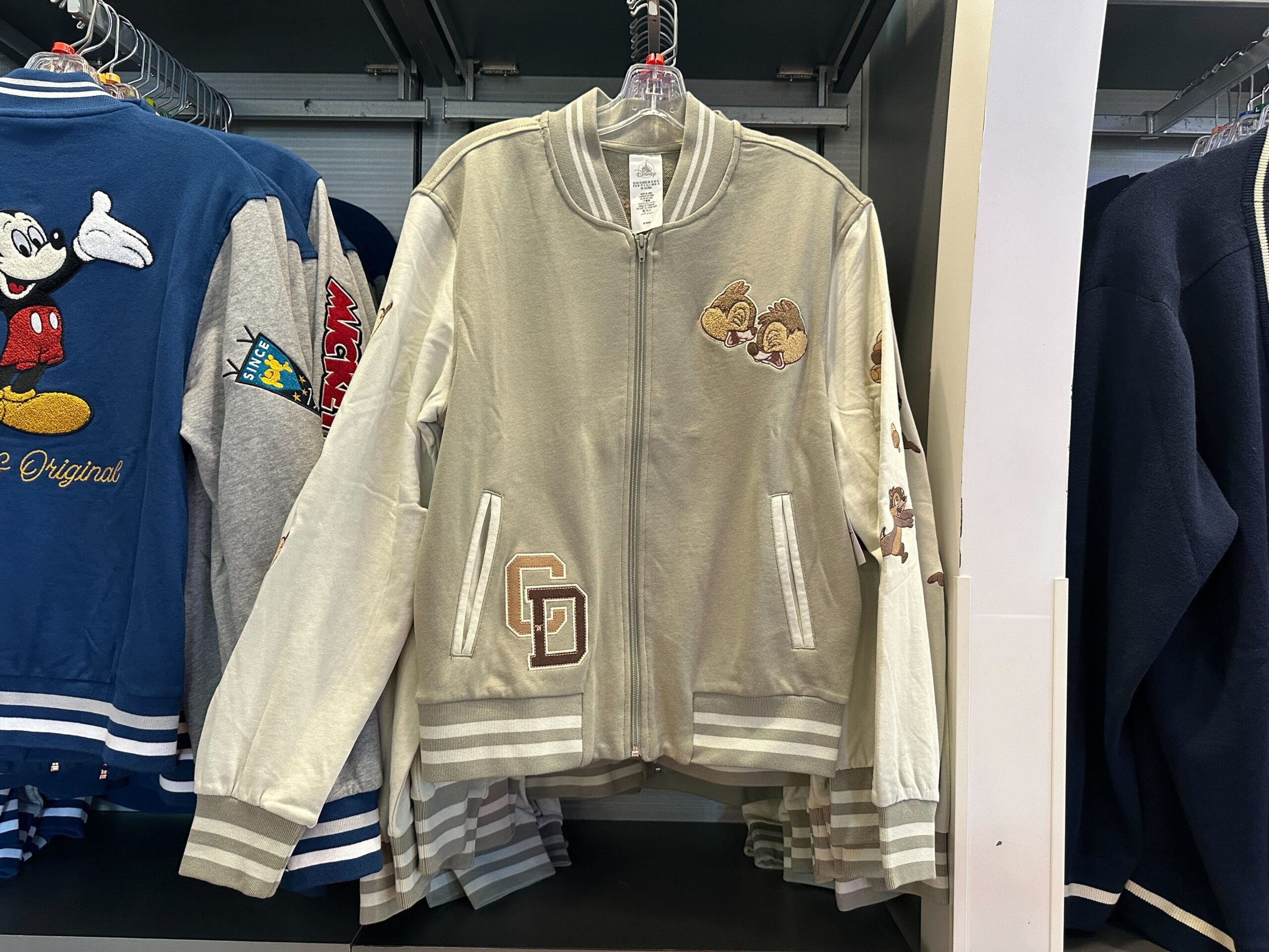 Light gray Disney letterman jacket with Chip 'n' Dale patches, hanging among other jackets in a store.