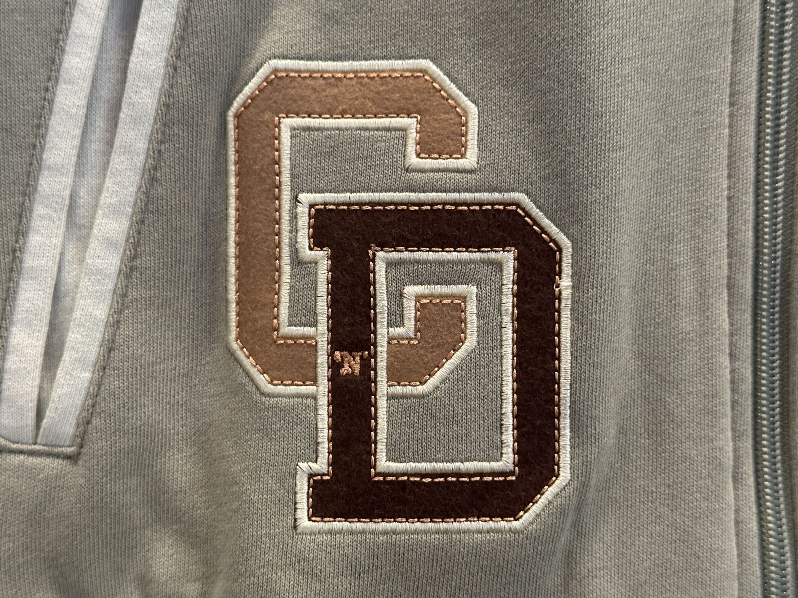 Close-up of Disney Letterman Jackets: embroidered "G" in tan, "D" in dark brown on gray fabric, with zipper and stripes.