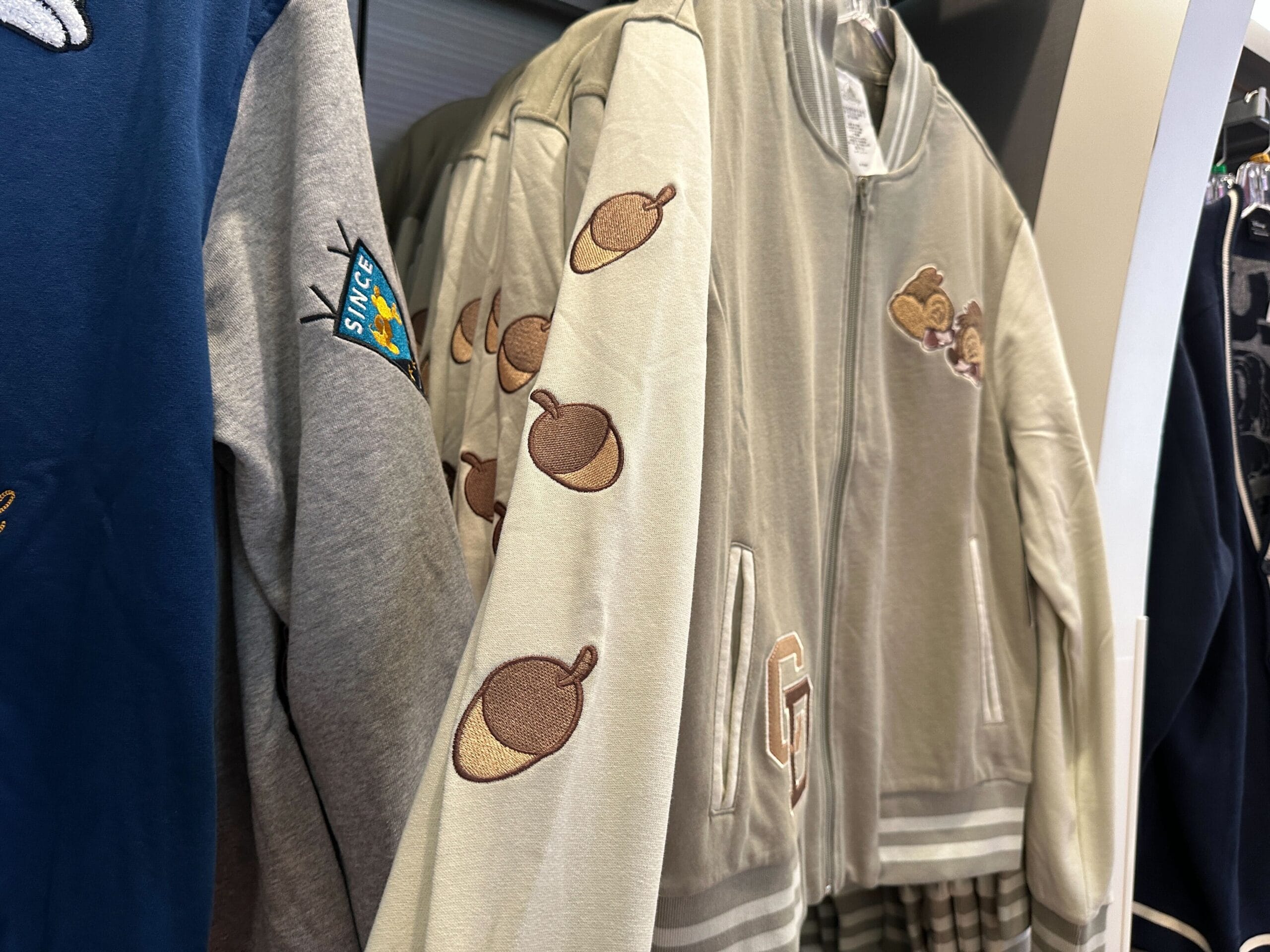Beige Disney Letterman Jacket with drumstick sleeves and bear patches, hanging on a clothing rack.