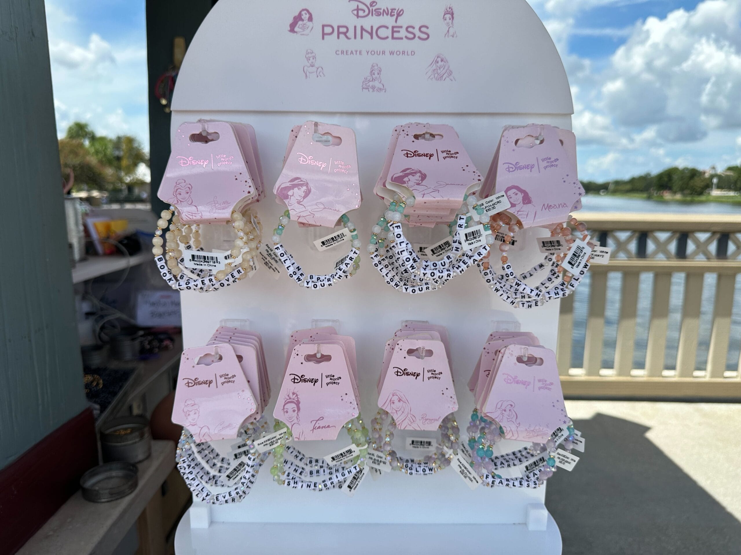 Display of Disney Princess beaded bracelets on a white stand, outdoors near a fence and water.