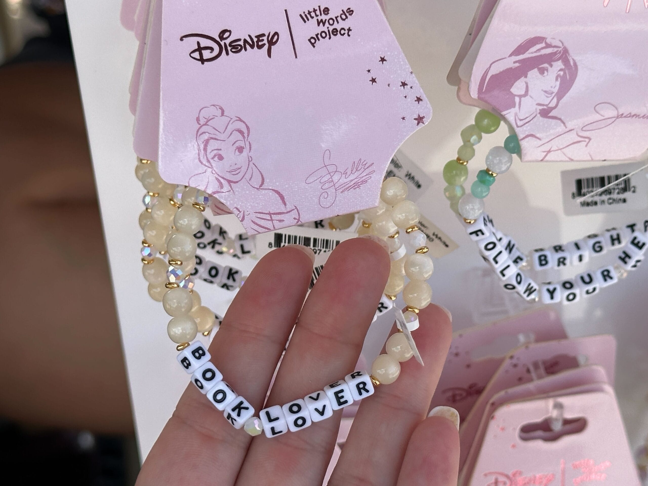 Hand holding a beaded bracelet that spells "BOOK LOVER," displayed among Disney princess-themed jewelry.