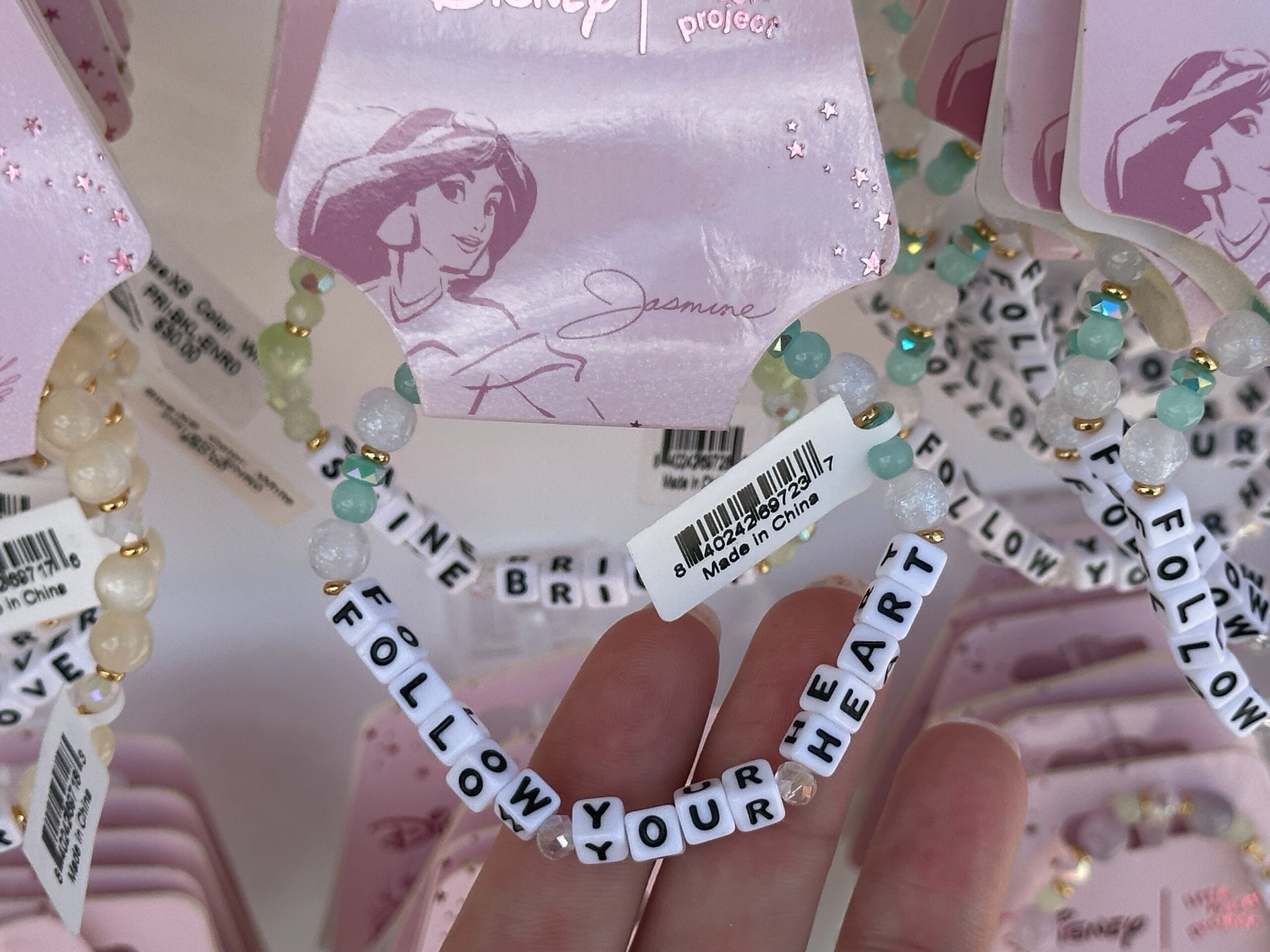 A hand holds a beaded bracelet reading "FOLLOW YOUR HEART" among similar bracelets on a store display.
