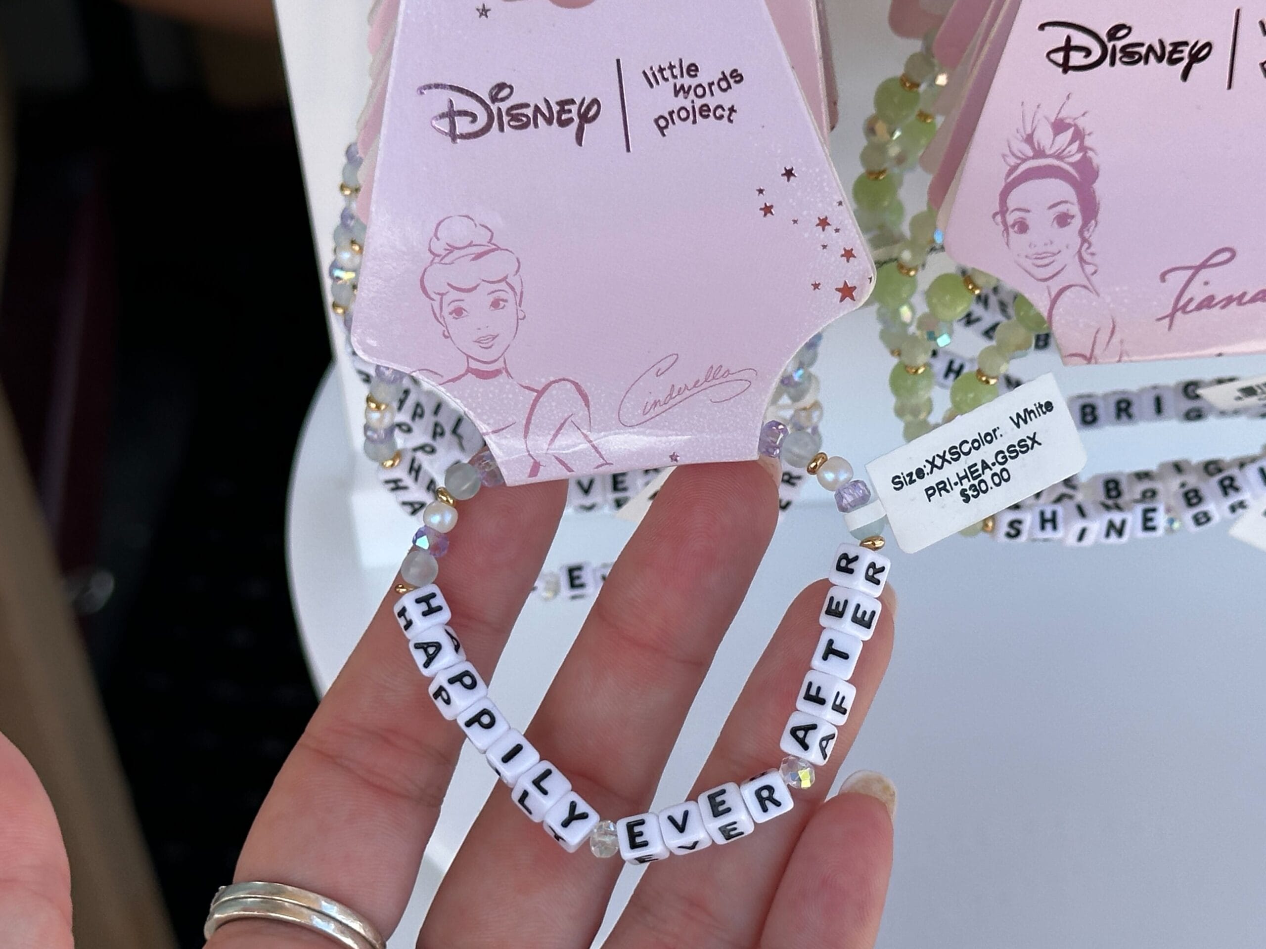 Hand holding a beaded bracelet with white letter beads spelling "HAPPILY EVER AFTER" and Disney princess tag attached.
