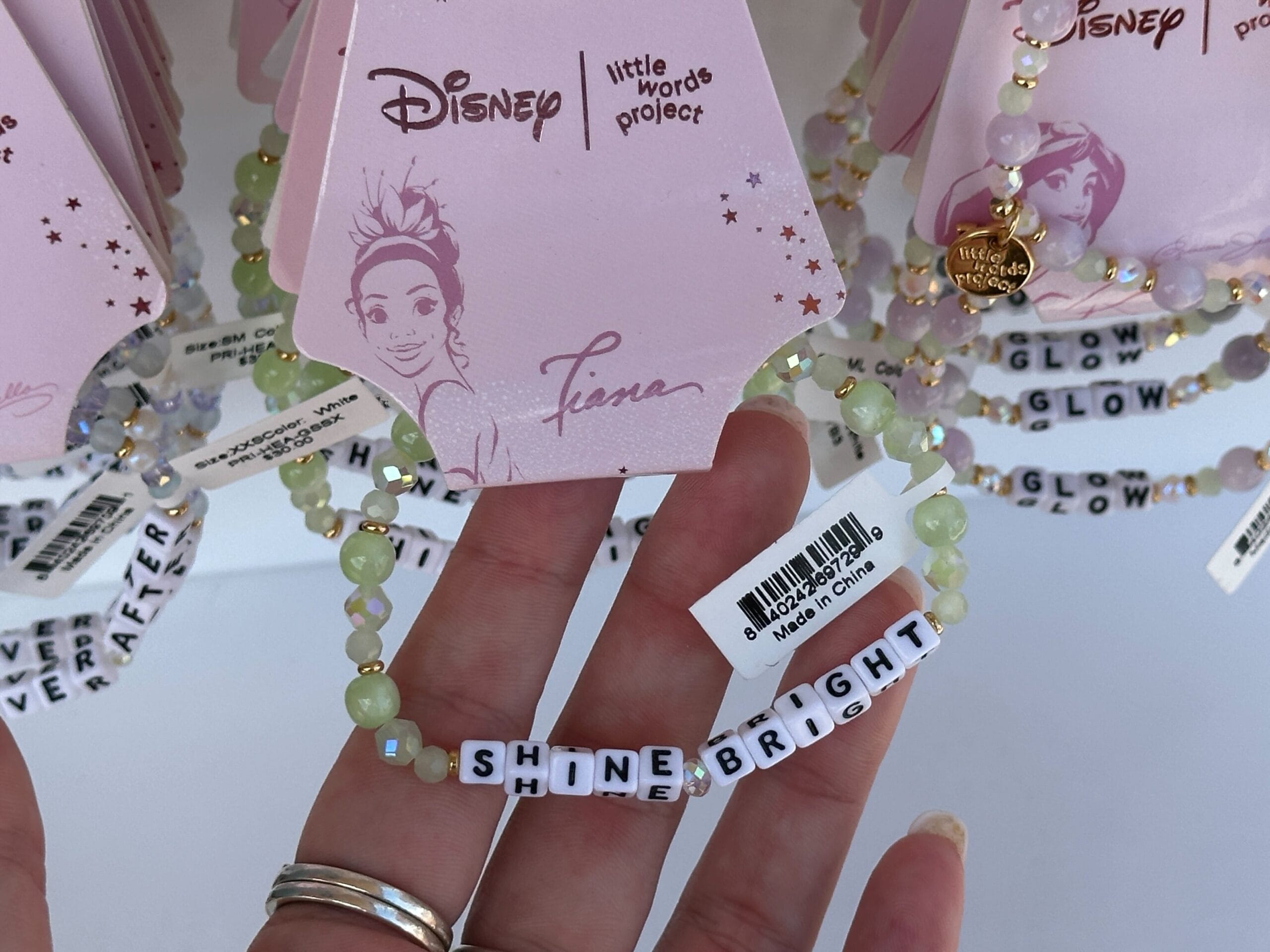 A hand holds a beaded bracelet with letter beads spelling "SHINE BRIGHT" under Disney Princess Tiana packaging.