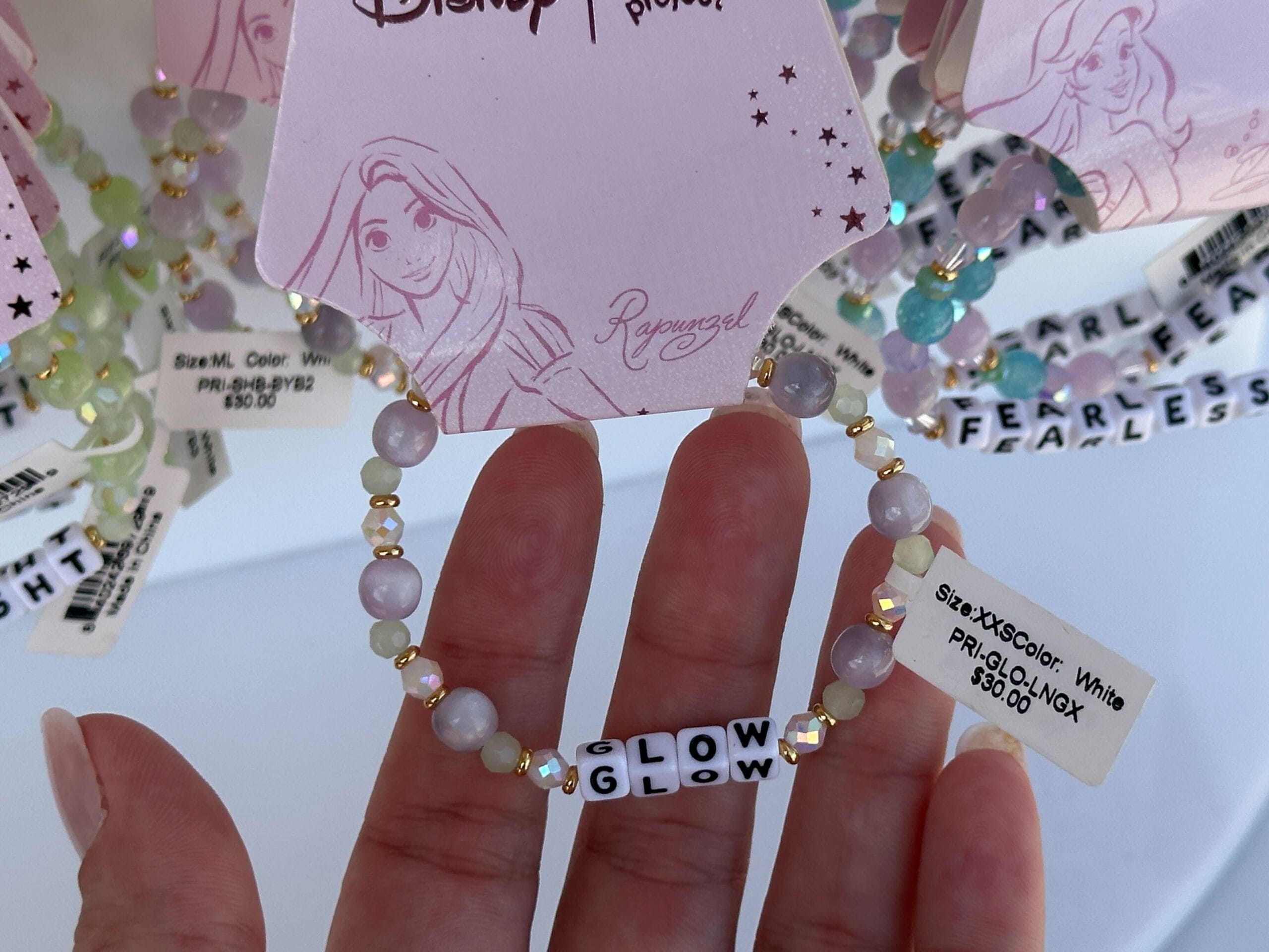 Hand holding a beaded bracelet with "GLOW GLOW" word beads, displayed on a Rapunzel-themed Disney tag.