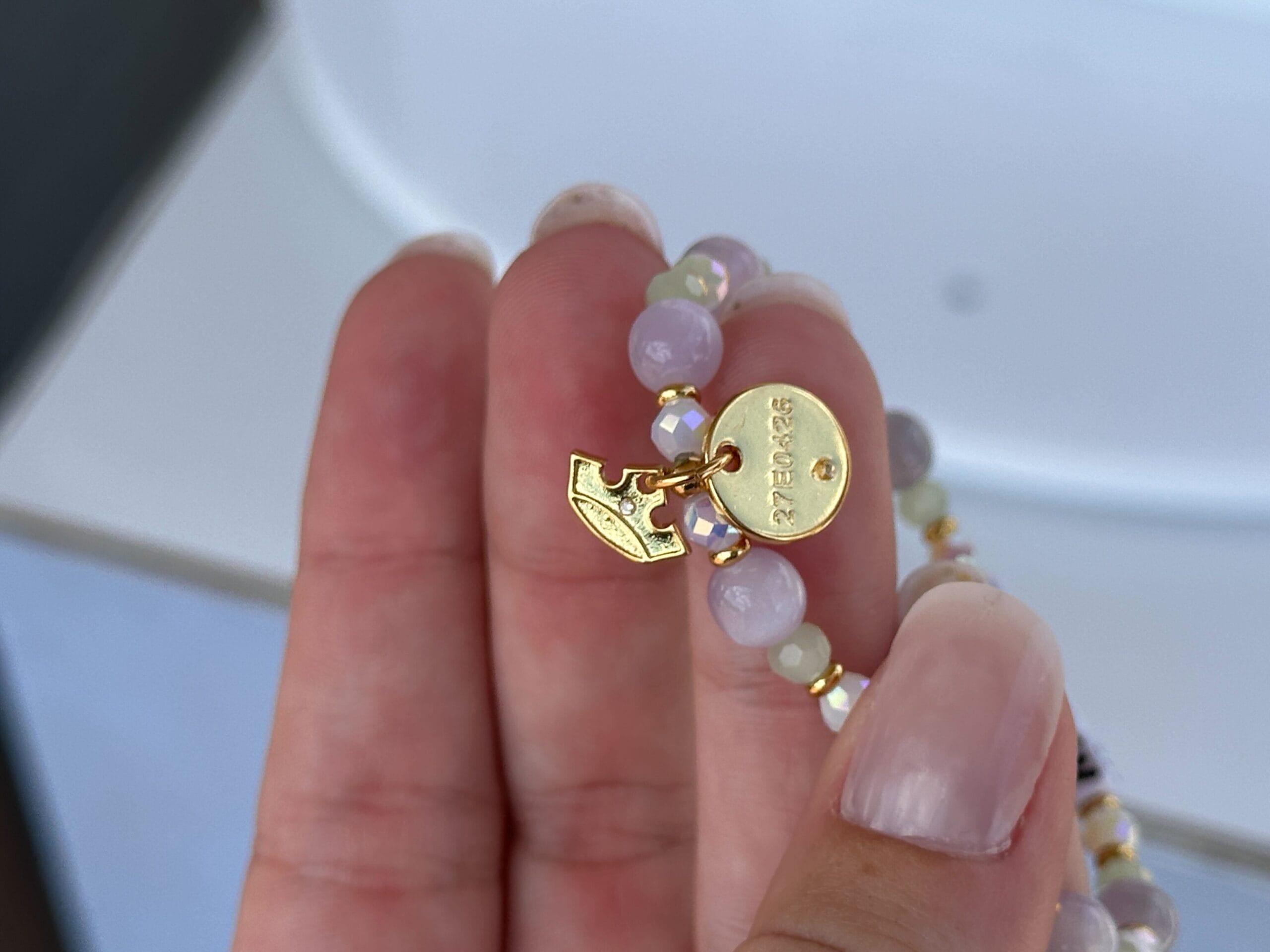 A hand holds a beaded bracelet with a gold oval tag engraved "GUESS" and a gold arrow charm.