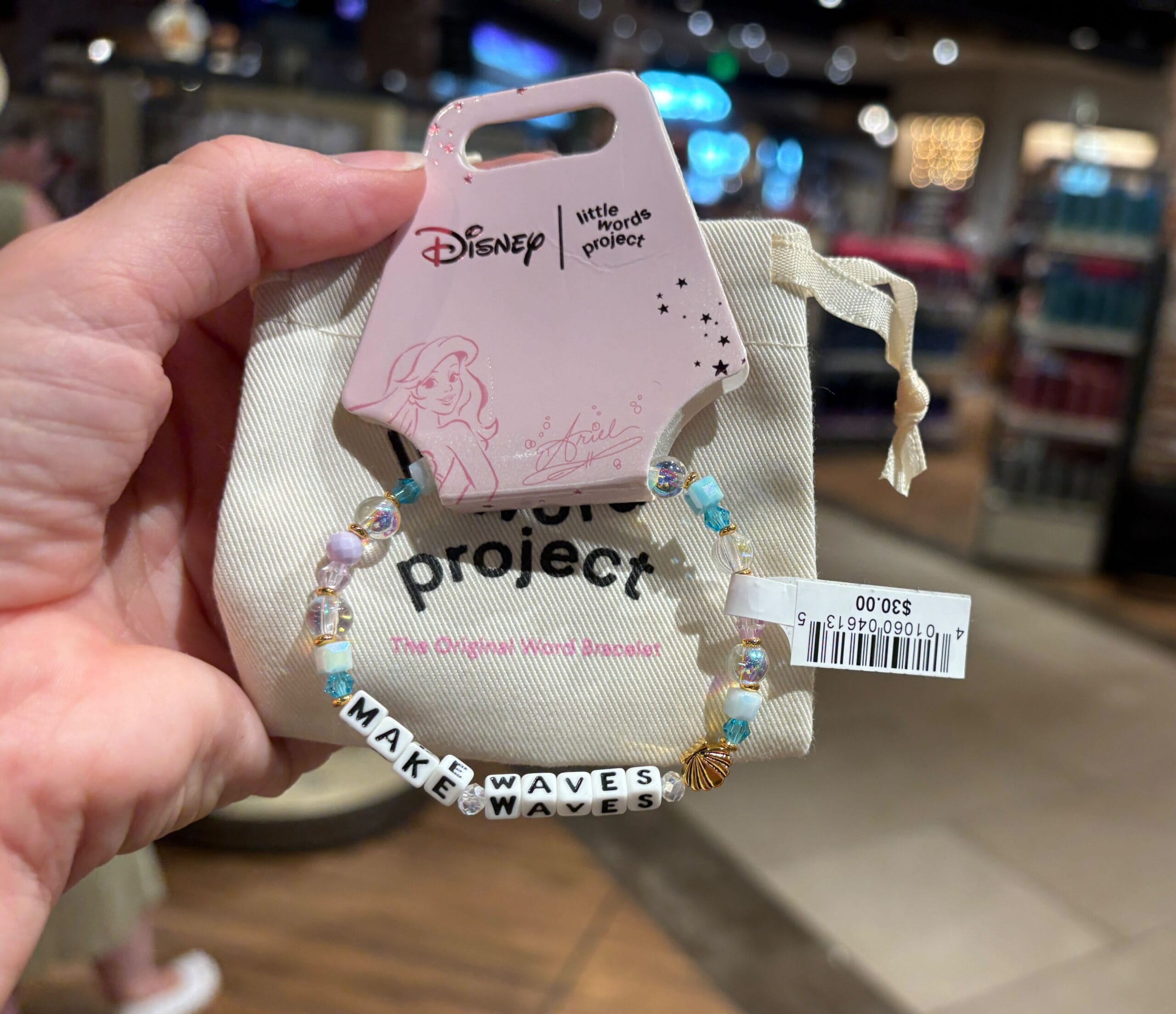 A hand holds a Disney "MAKE WAVES" beaded bracelet with Ariel on the tag in a store.