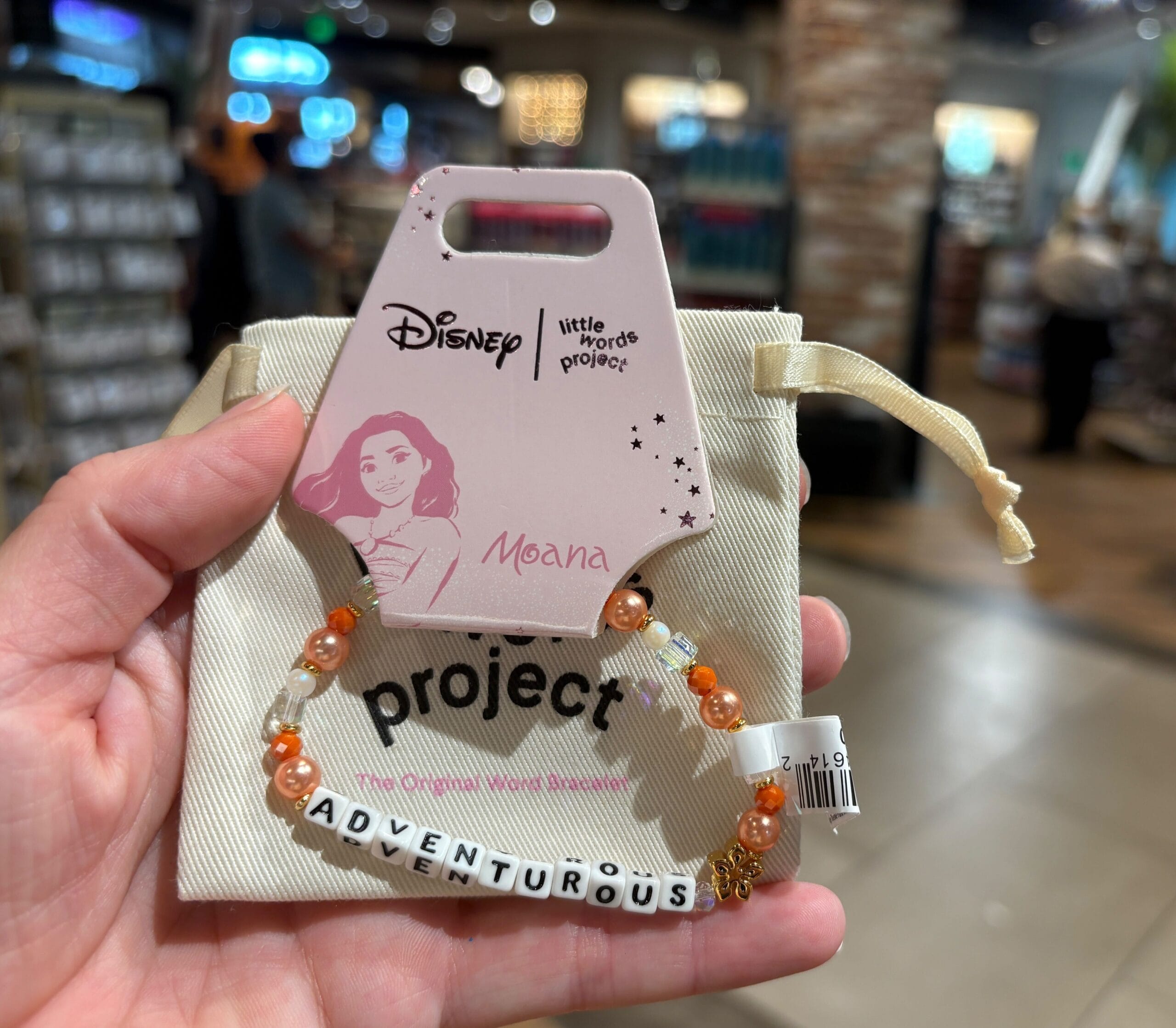A hand holds a Moana-themed beaded bracelet spelling "ADVENTUROUS" in a store.