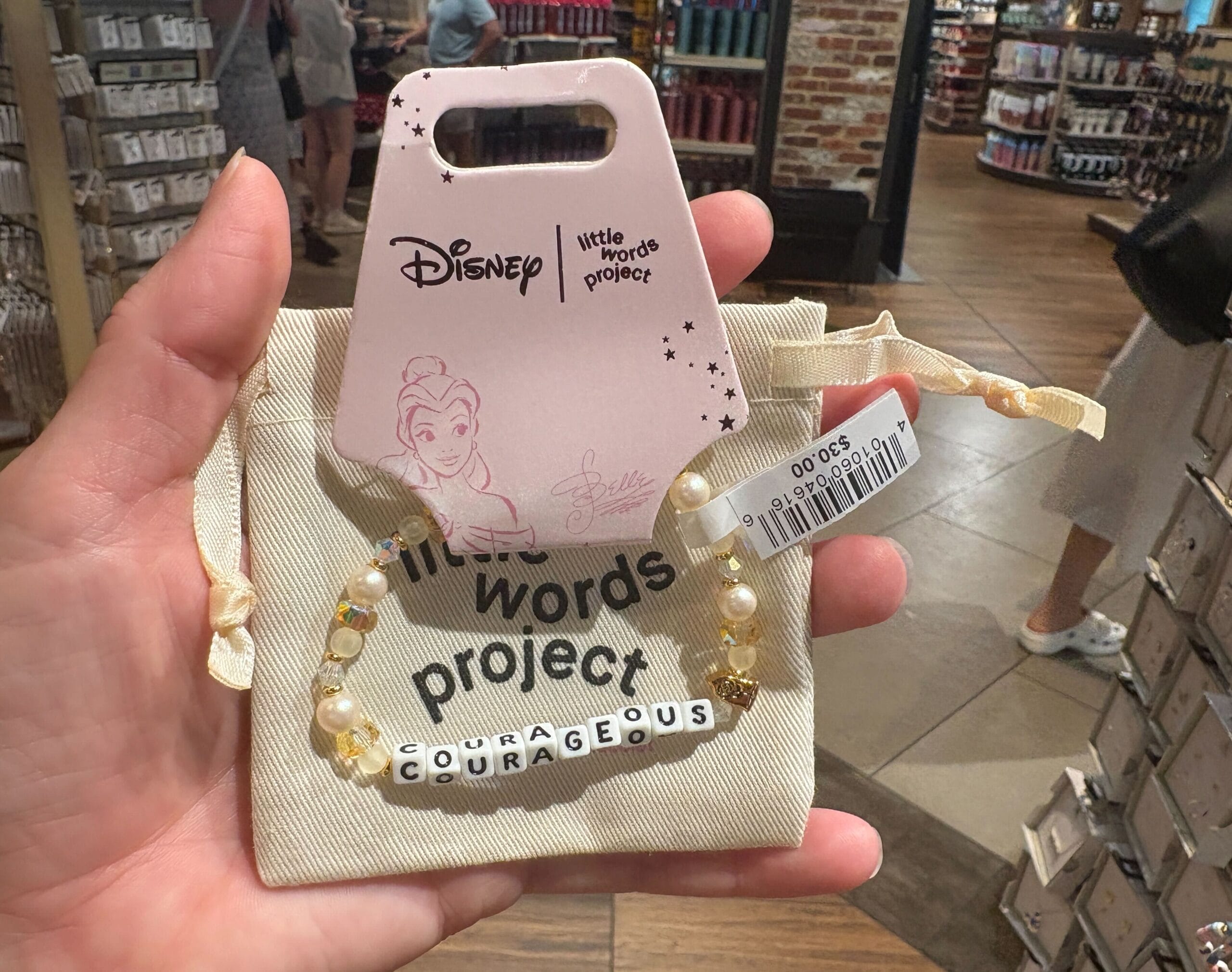 A hand holds a Disney "Courageous" beaded bracelet with Belle on pink tag, inside a store.