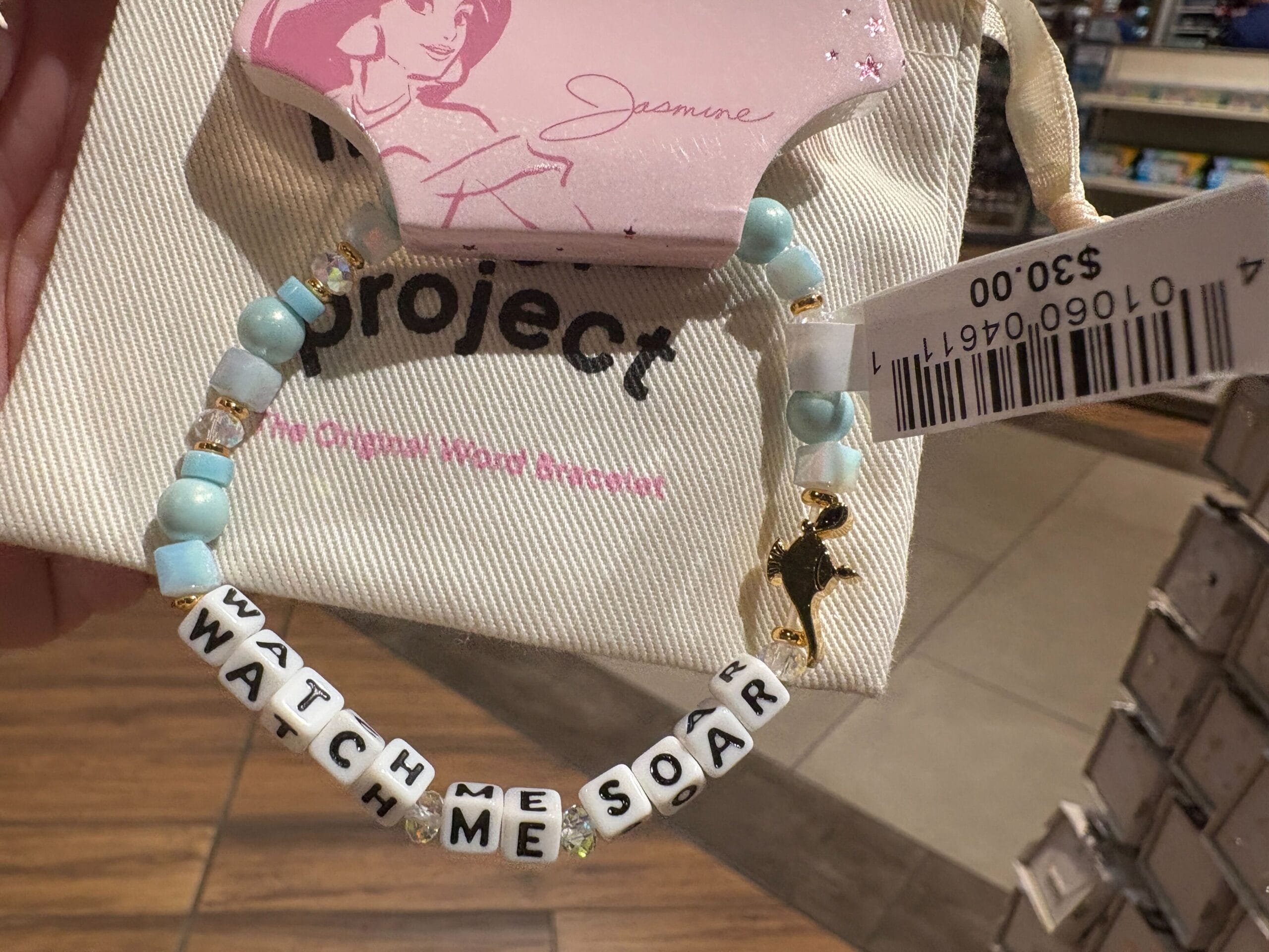 A beaded bracelet spells "WATCH ME SOAR" with a gold bird charm, displayed in a store with a $3 price tag.