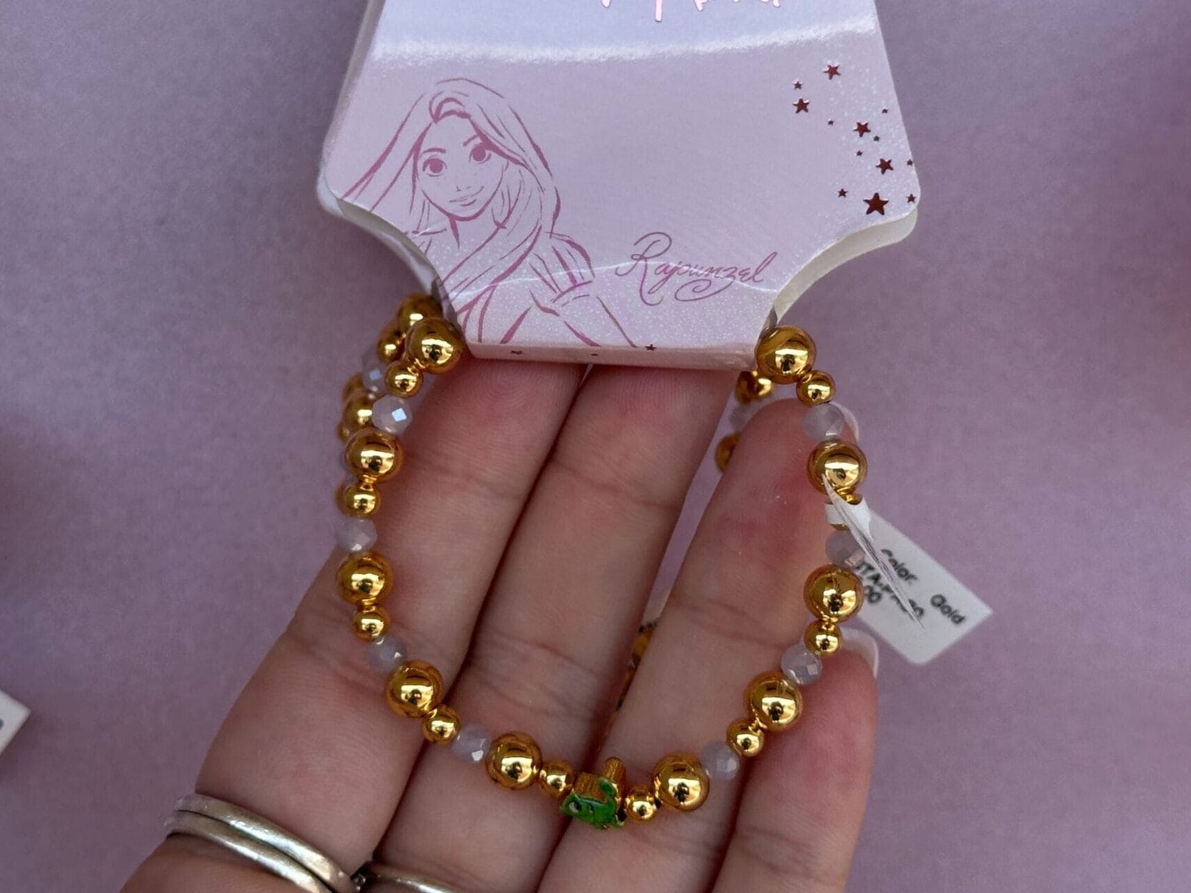 Hand holding a Disney Rapunzel bracelet with gold beads and a green accent, displayed on pink packaging.