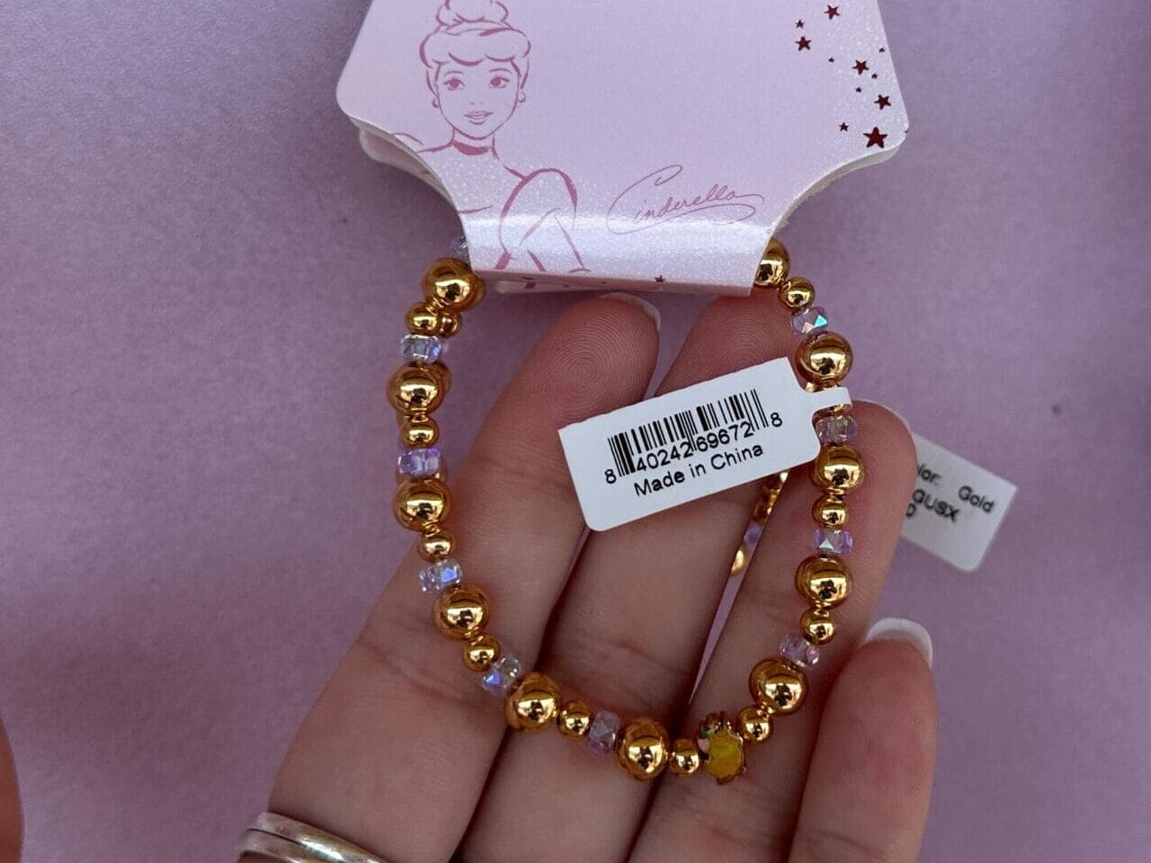A hand holds a gold beaded Disney Cinderella bracelet against a pink display with other beaded bracelets.