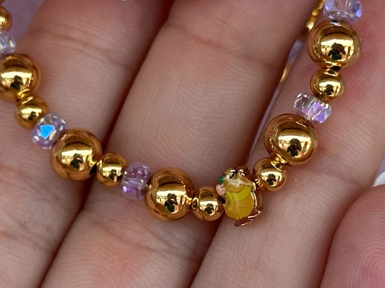 A hand holds a gold beaded bracelet with purple accents and a yellow bead, against a pink background.