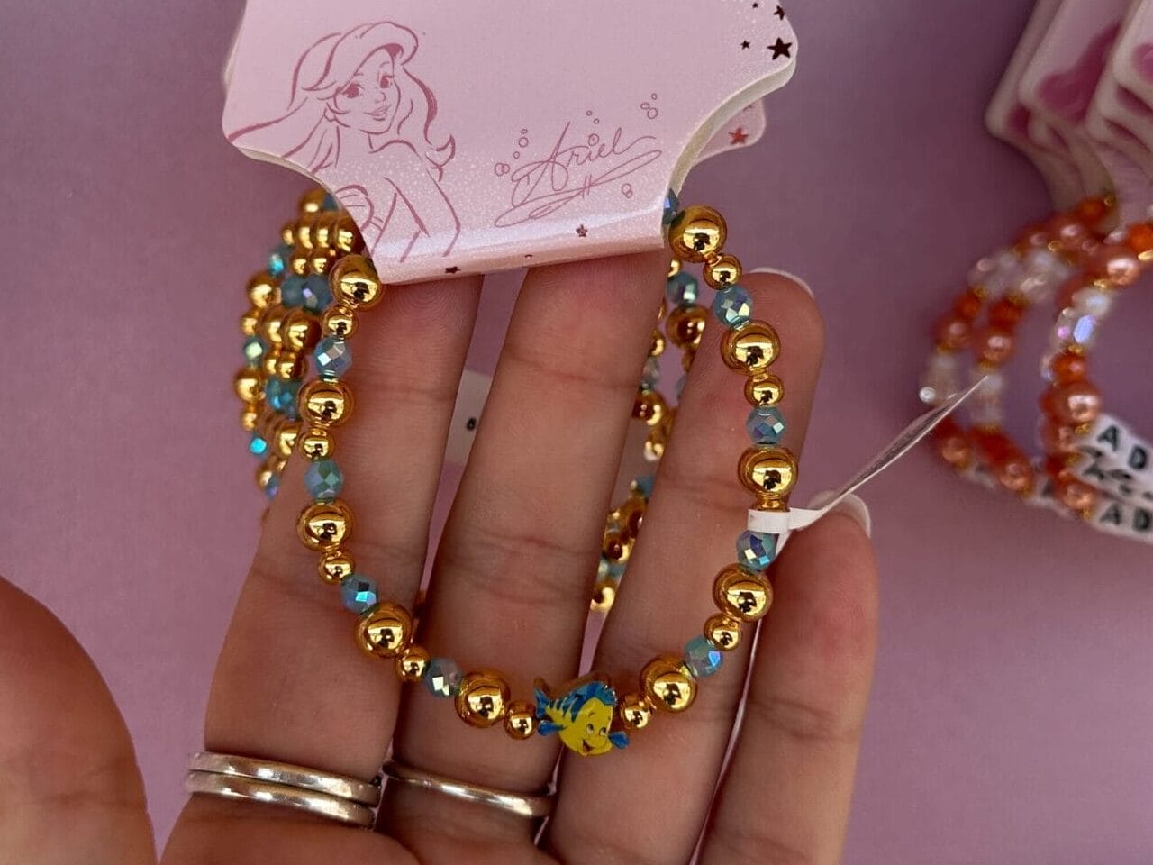 A hand holds up Disney Princess beaded hoop earrings against a display of similar earrings on a pink background.