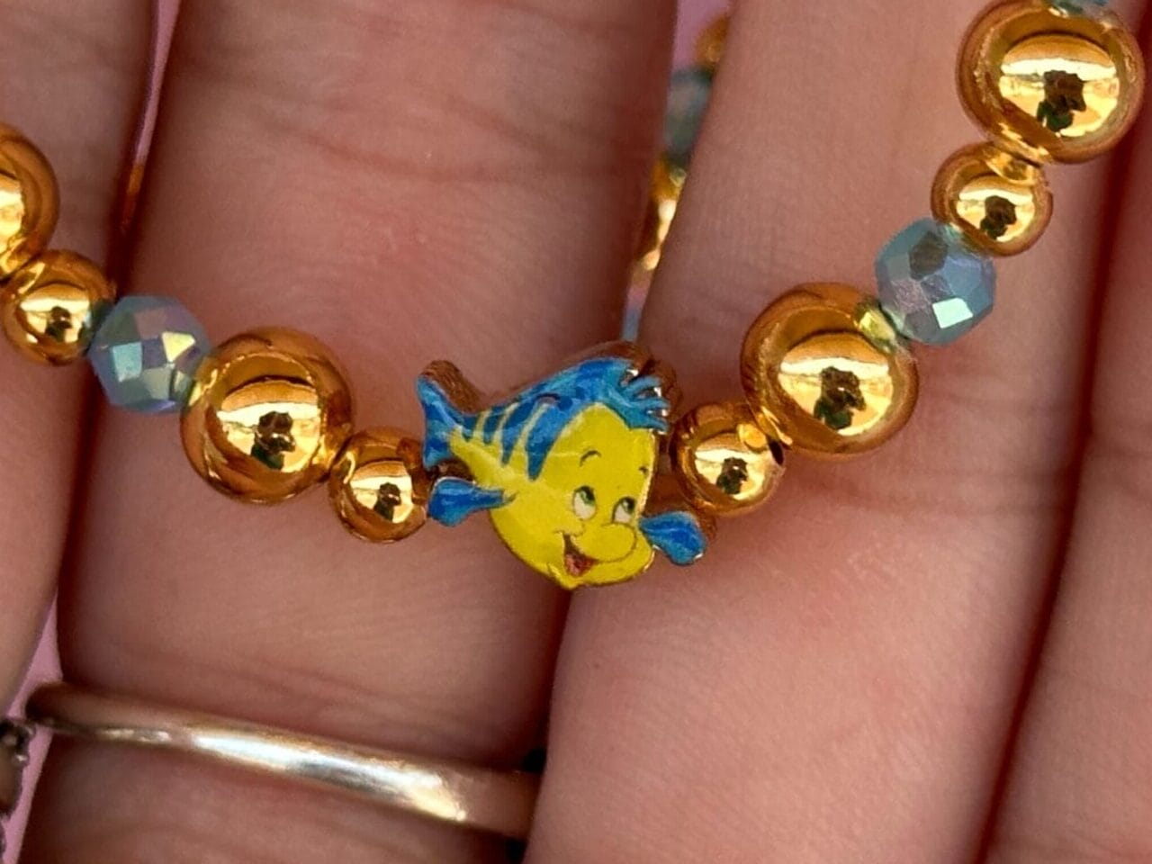A hand holds a beaded bracelet with gold beads and a small blue and yellow fish charm.
