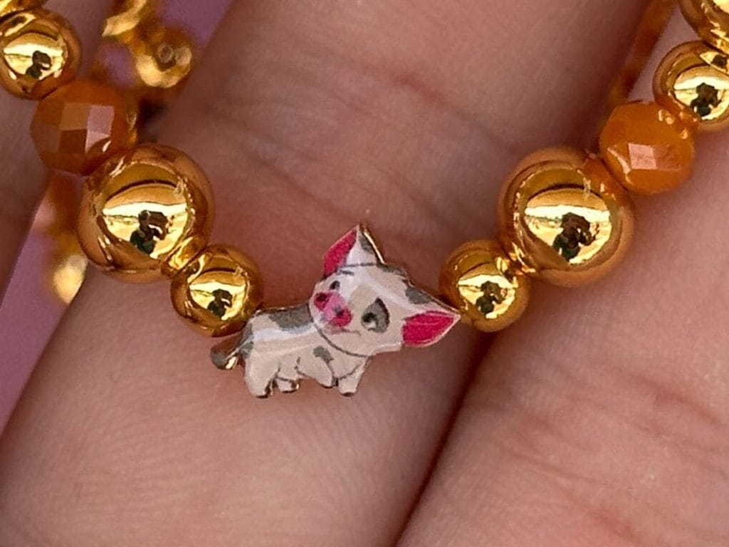 A hand holding a beaded bracelet with gold and orange beads, featuring a pig charm in the center.