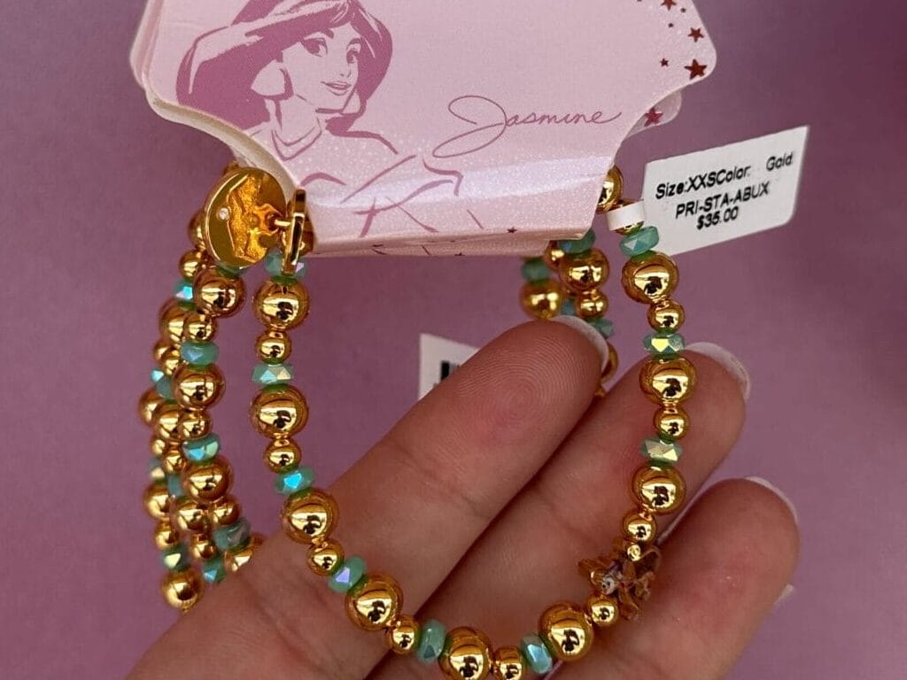 A hand holds a Disney Princess Jasmine beaded bracelet set with gold and turquoise beads against a pink background.