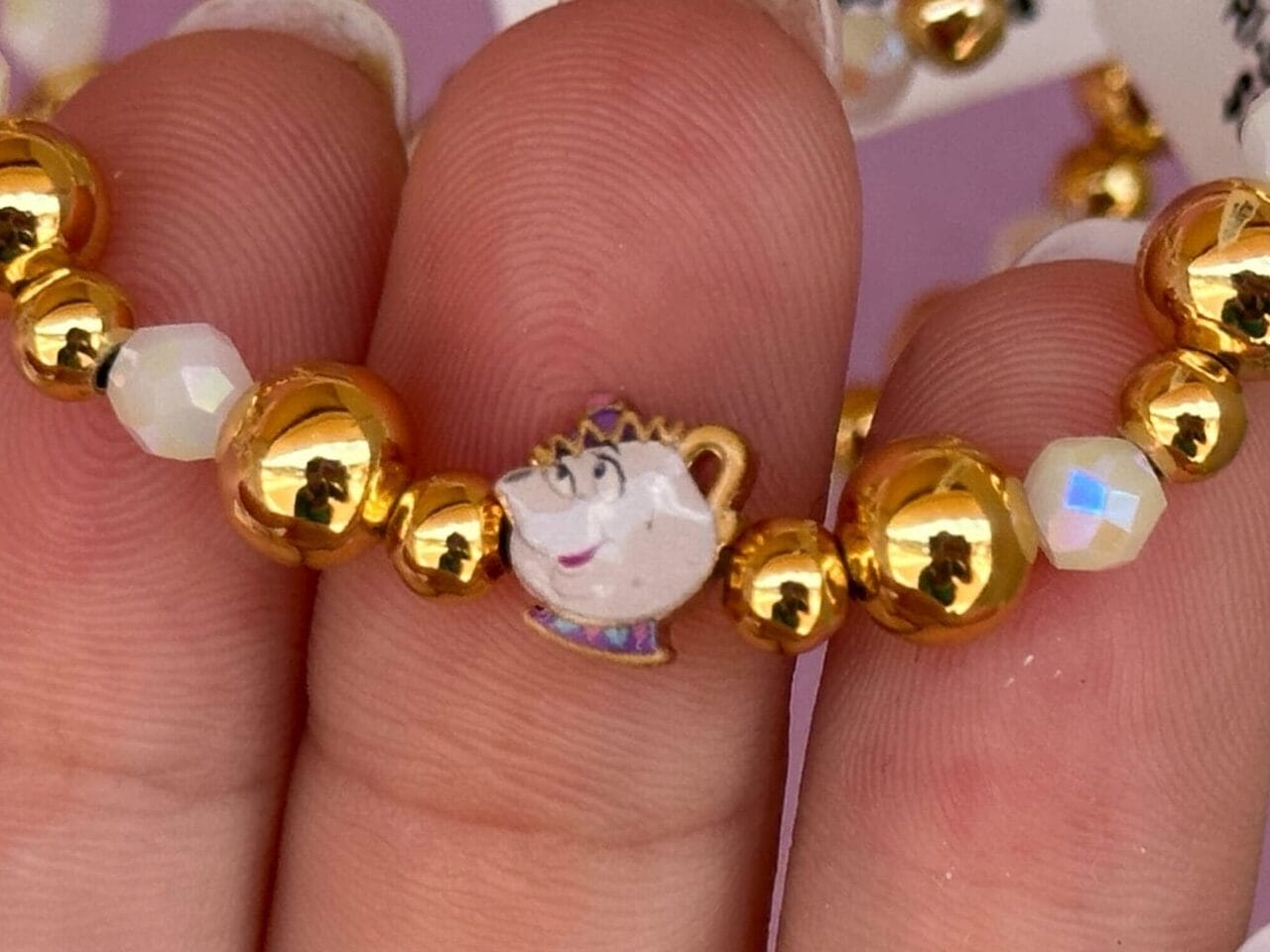 Hand holding a gold and white beaded bracelet with a small Chip the teacup charm from Beauty and the Beast.