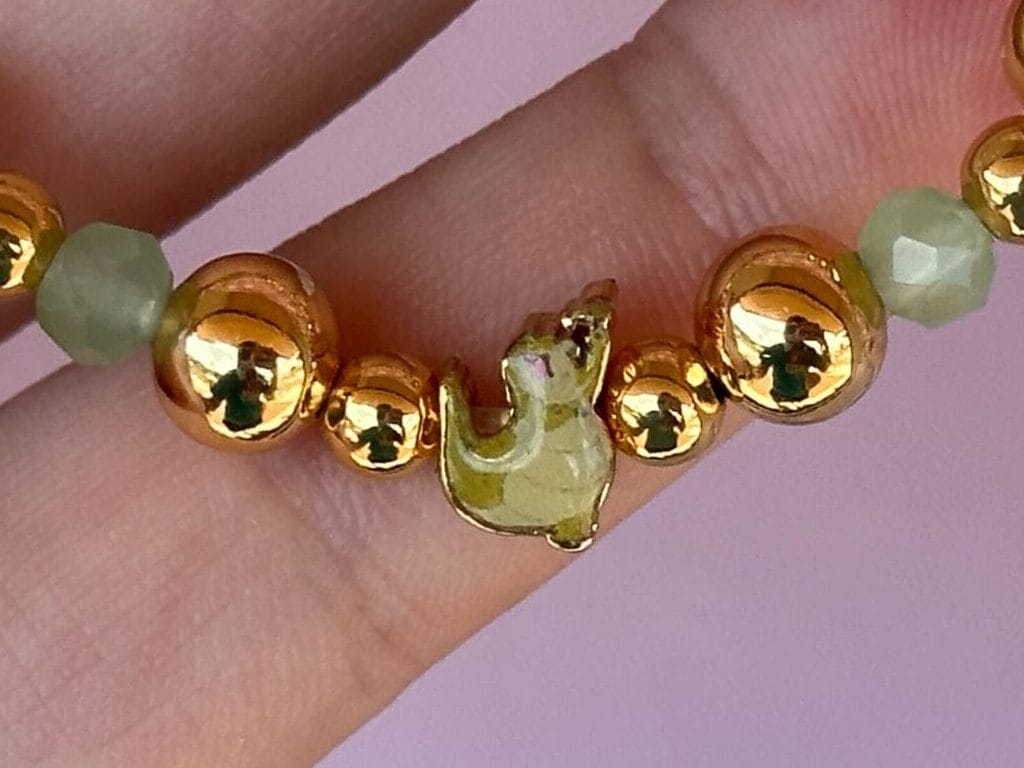 A hand holds a beaded bracelet with gold and light green beads against a pink background.