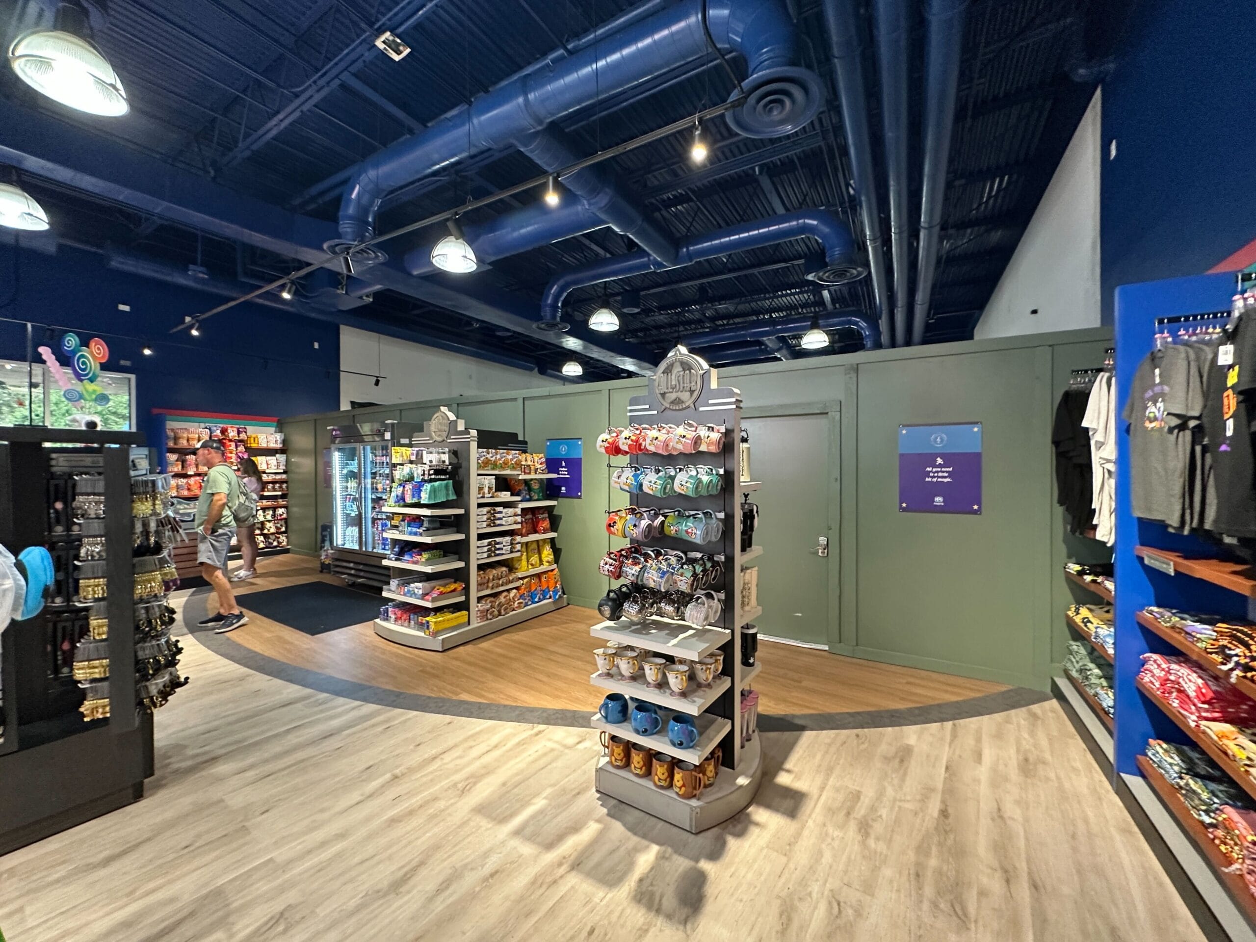 A retail store interior with snacks, drinks, and souvenirs on shelves; two people browse near the back.