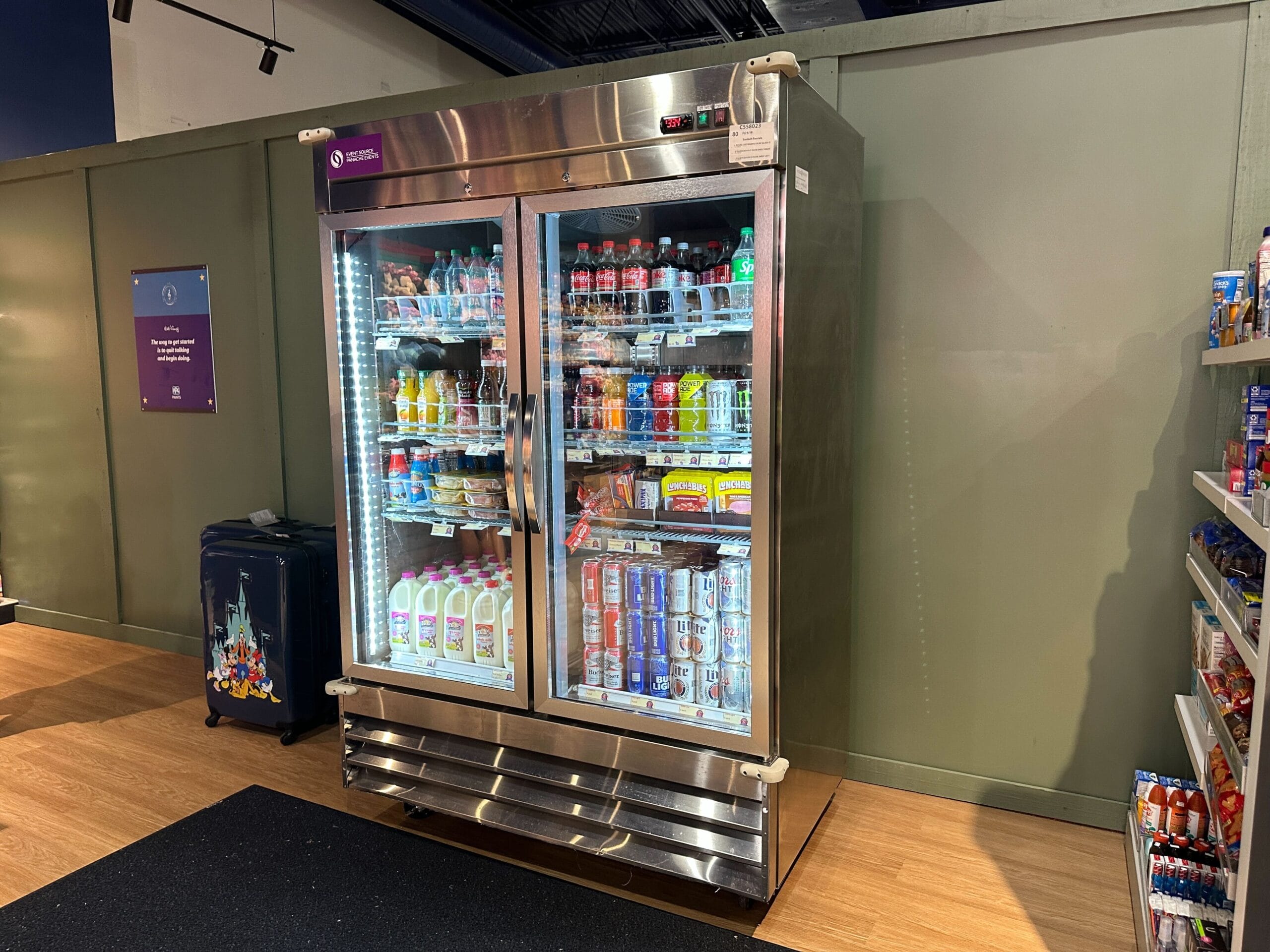 Double-door refrigerator filled with drinks like milk, soda, and juice, next to shelves of snacks in a store.