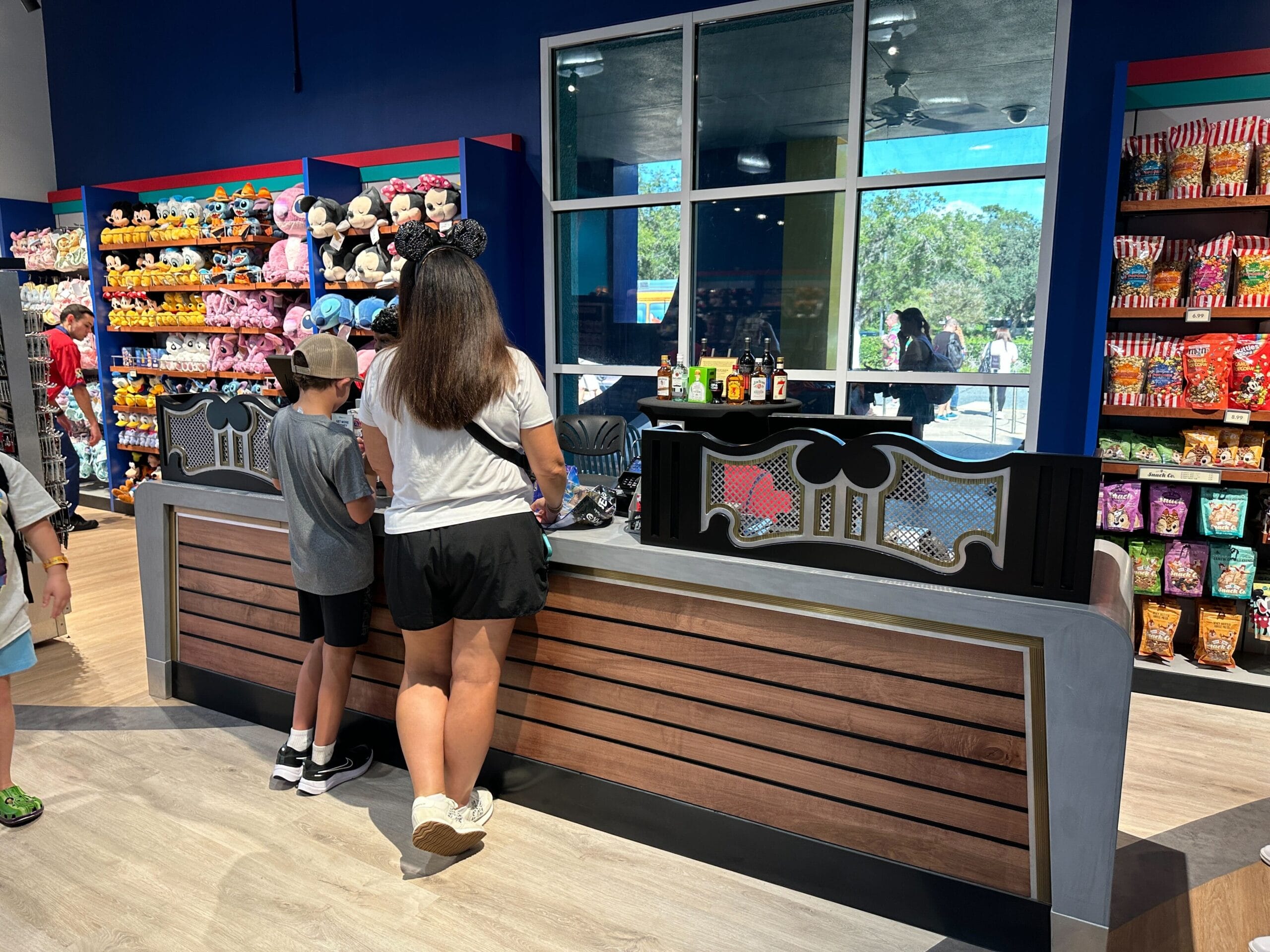 A woman and boy stand at a store counter with shelves of plush toys and snacks in the background.
