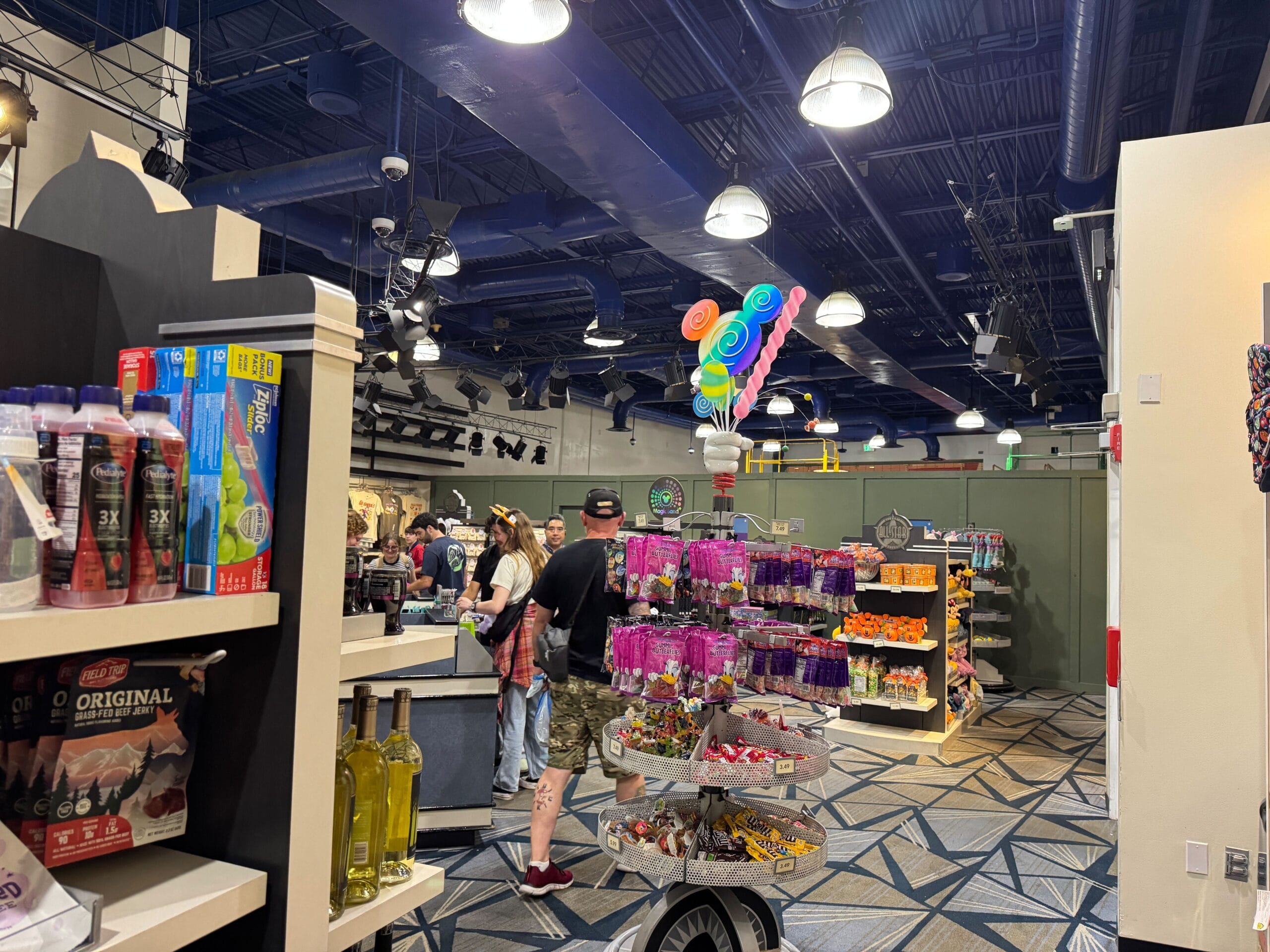 A store interior with people shopping, shelves of snacks and drinks, and a display of colorful lollipop balloons.