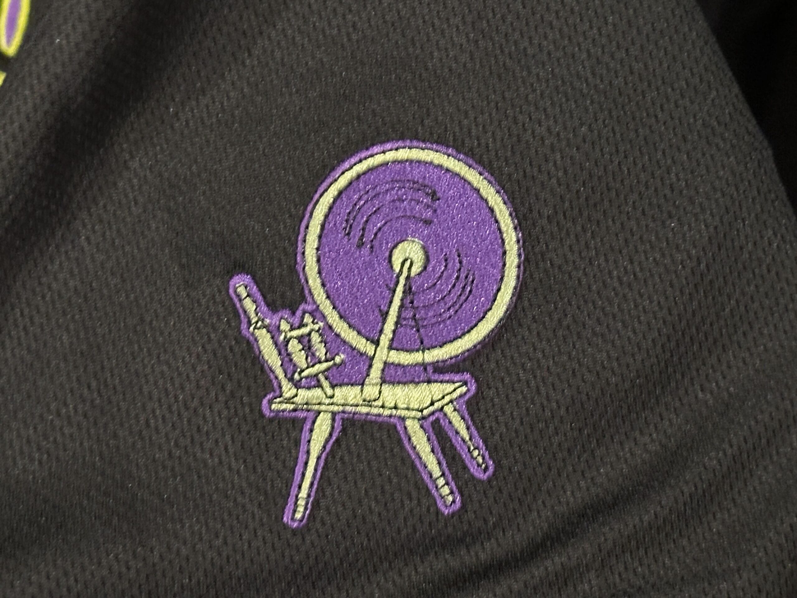 Purple and yellow embroidery of a spinning wheel on a chair, stitched on a black fabric background.