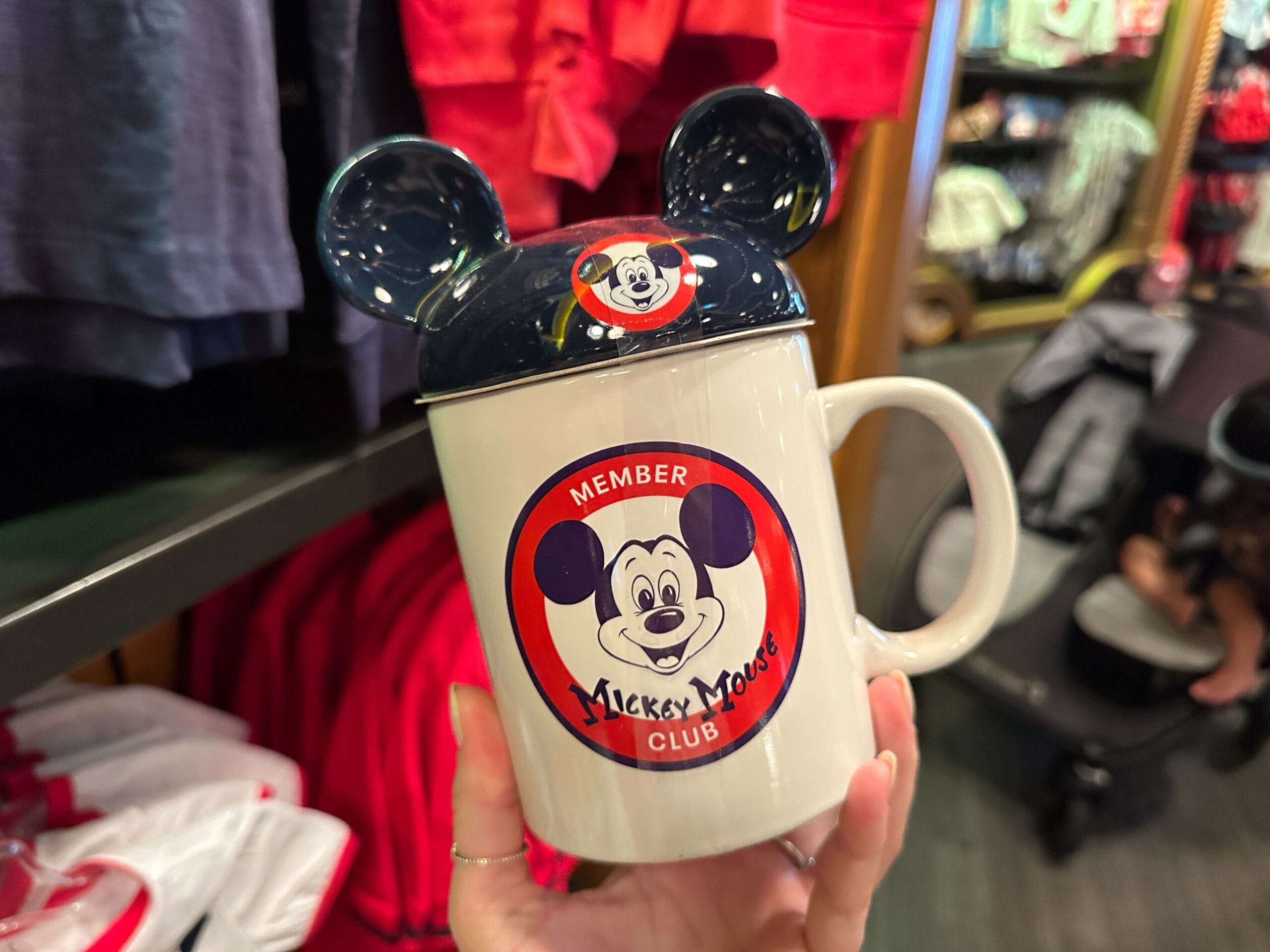 A hand holds a Mickey Mouse Club mug with a lid shaped like Mickey ears in a store.