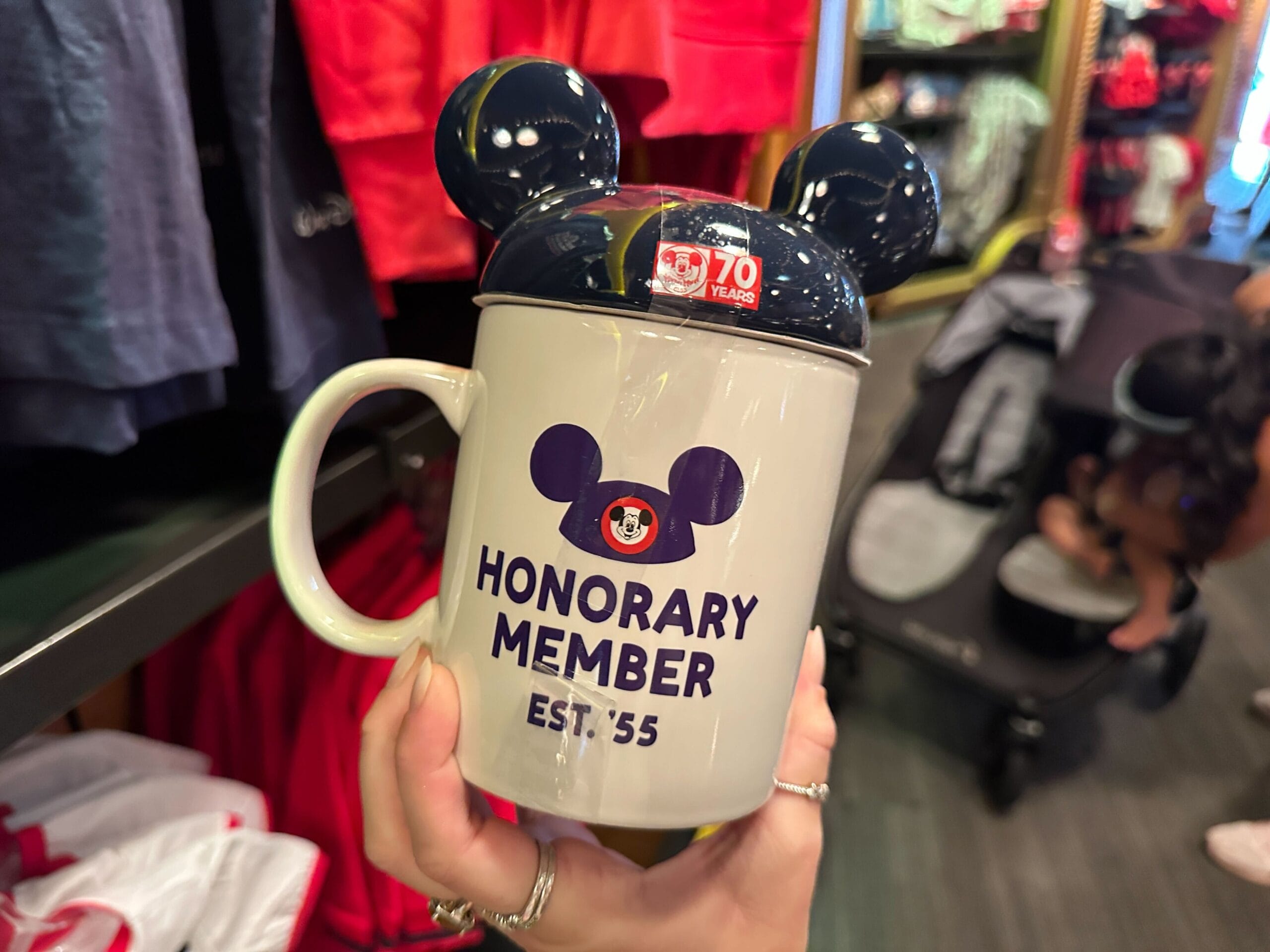 A mug with Mickey Mouse ears and "Honorary Member Est. '55" printed on it is held in a store.