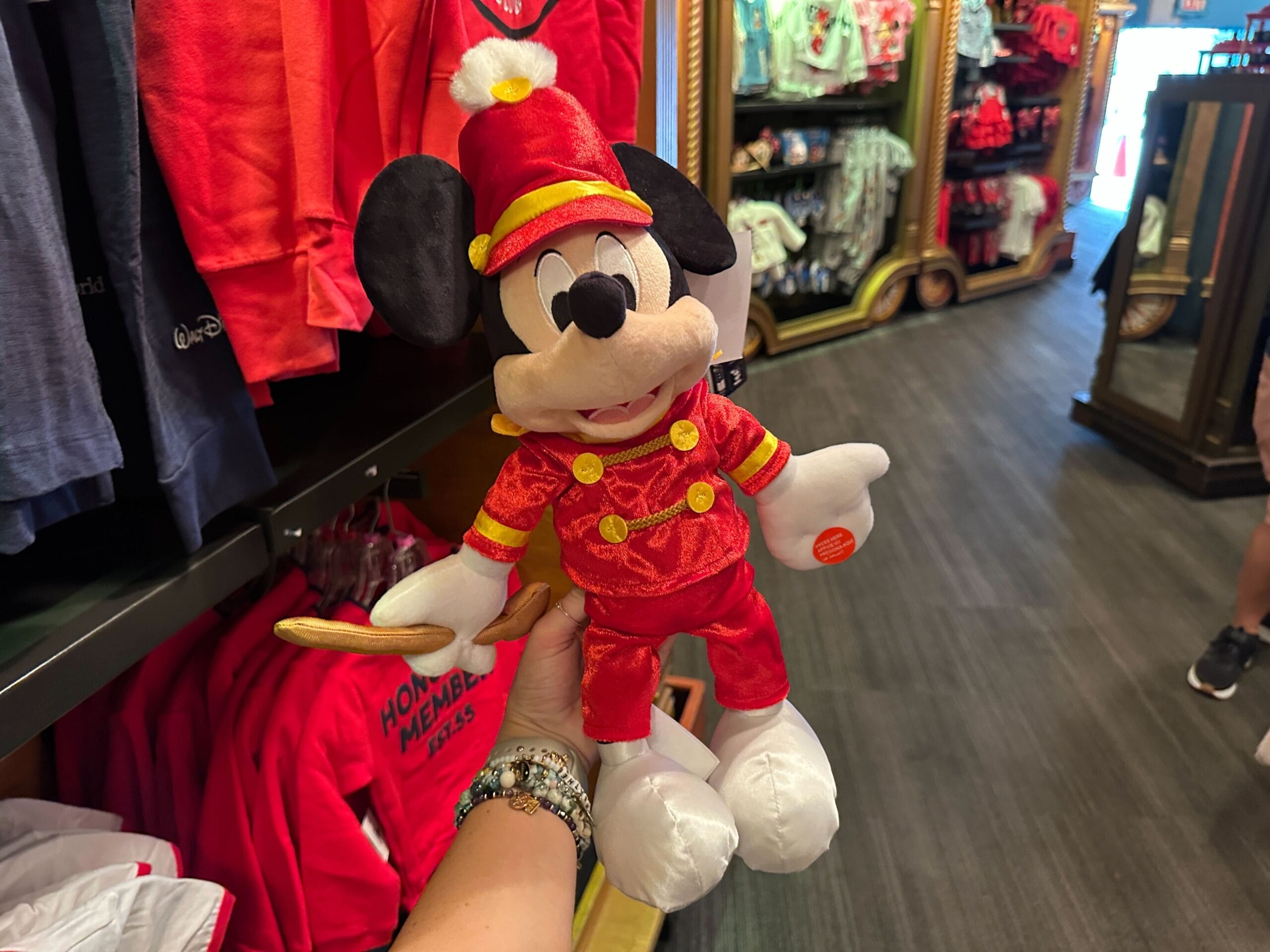 A hand holds a plush Mickey Mouse dressed as a red marching band leader in a store with colorful clothes in the background.