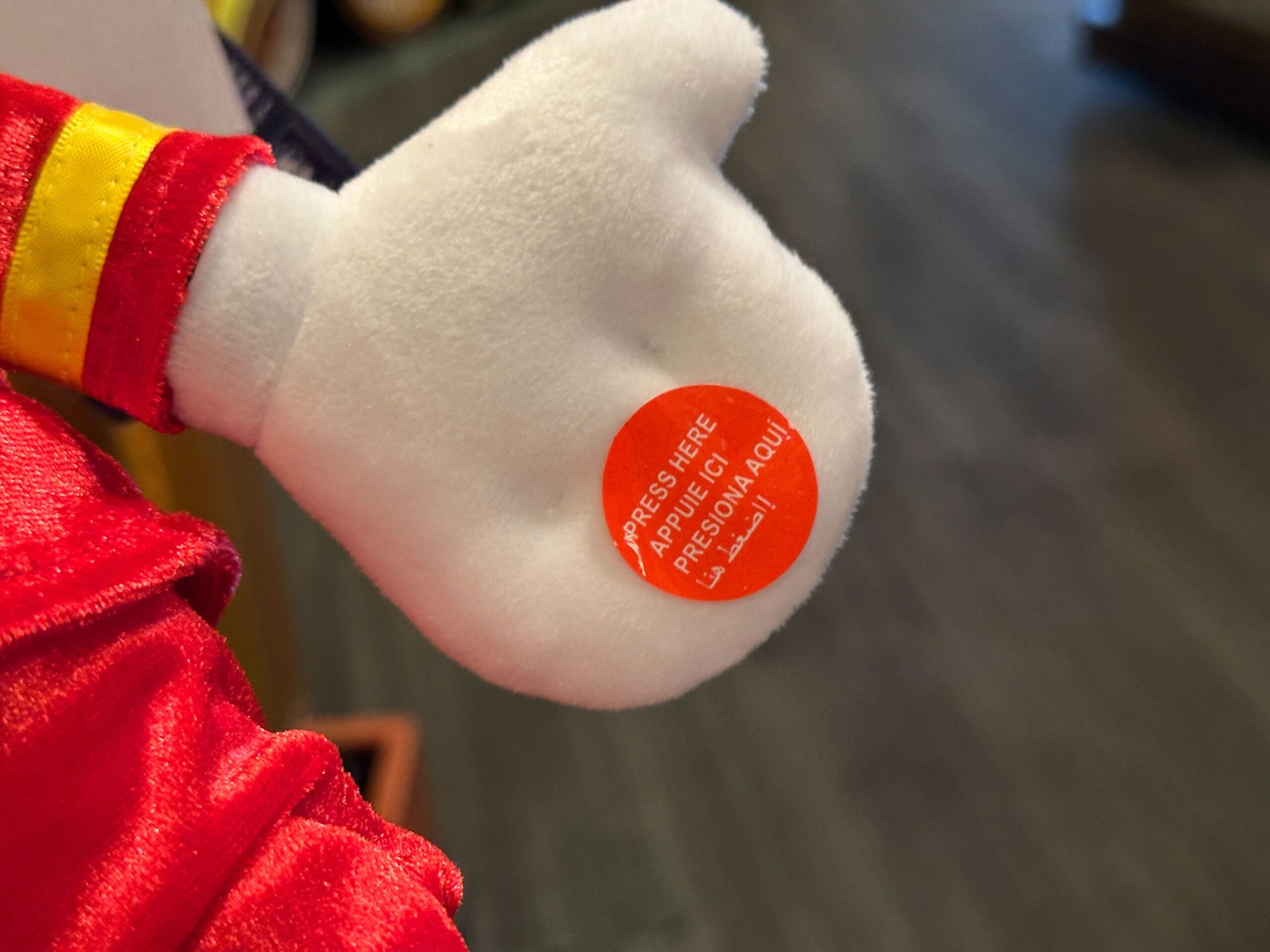 A plush character’s white-gloved hand with an orange sticker that says, "PRESS HERE / APRETAR AQUÍ.